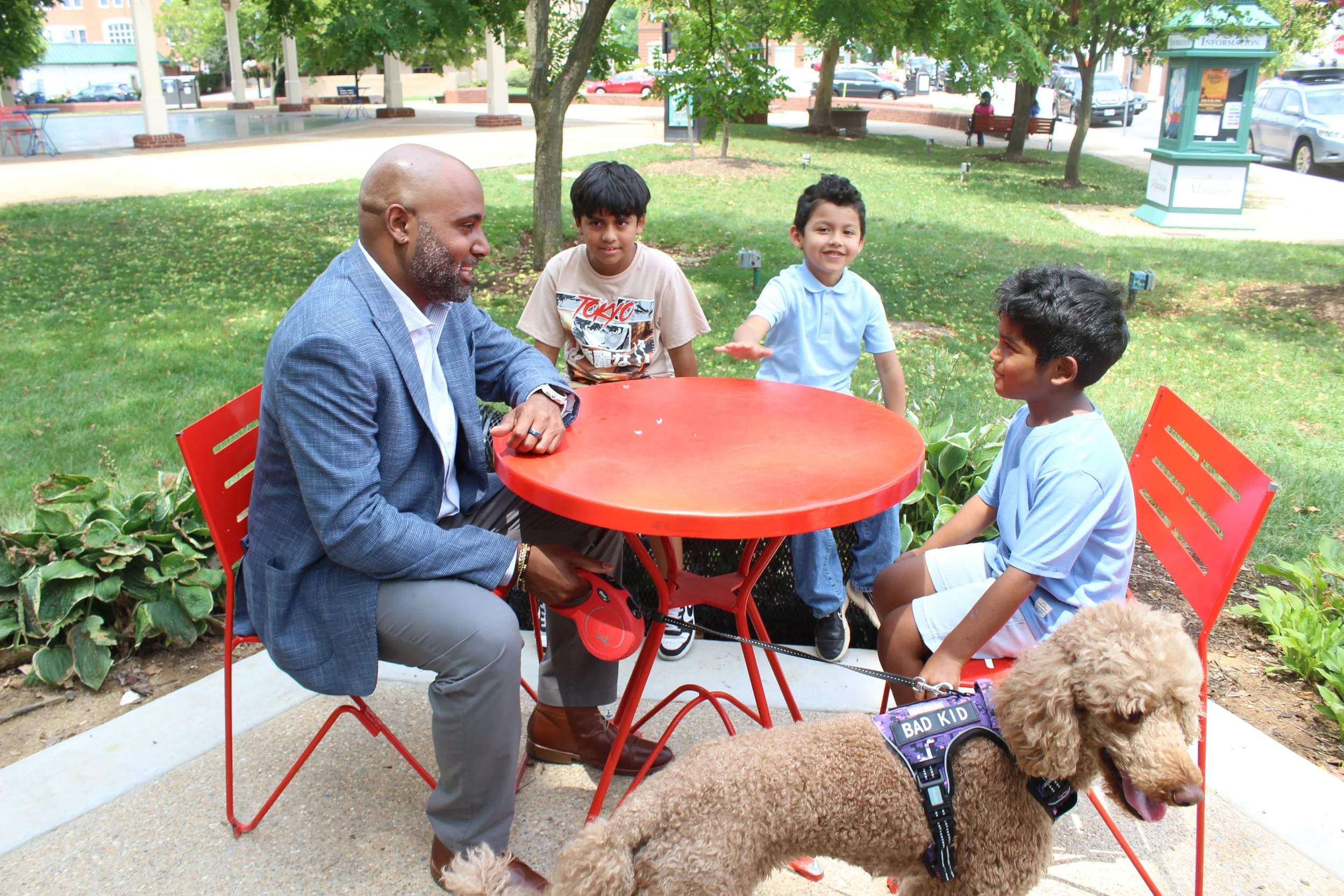 Four people, including a man and three boys, sitting outdoors at a red metal table in a park, with dogs around them, and trees and benches in the background.