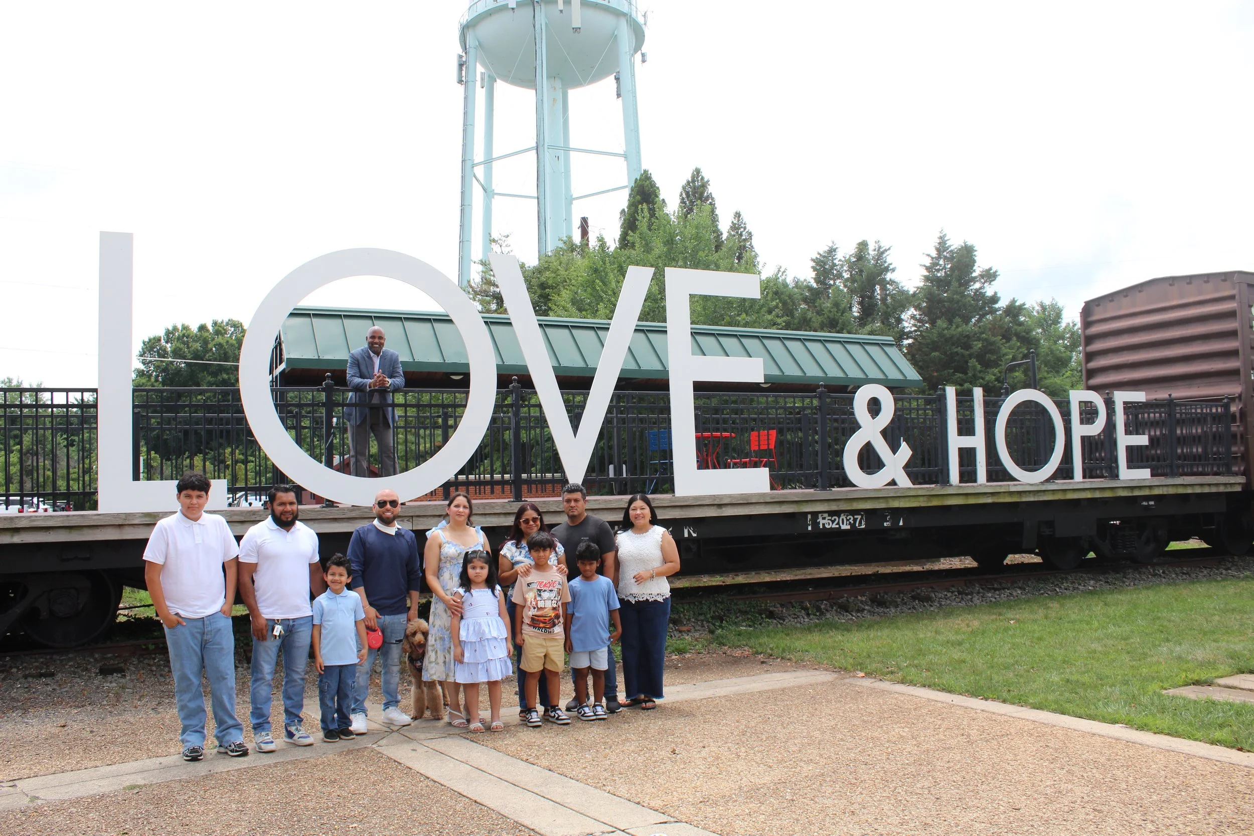 A group of people, including adults and children, standing in front of a large sign that says 'LOVE & HOPE' with a person standing on a platform behind the sign. The background features trees and a tall water tower.