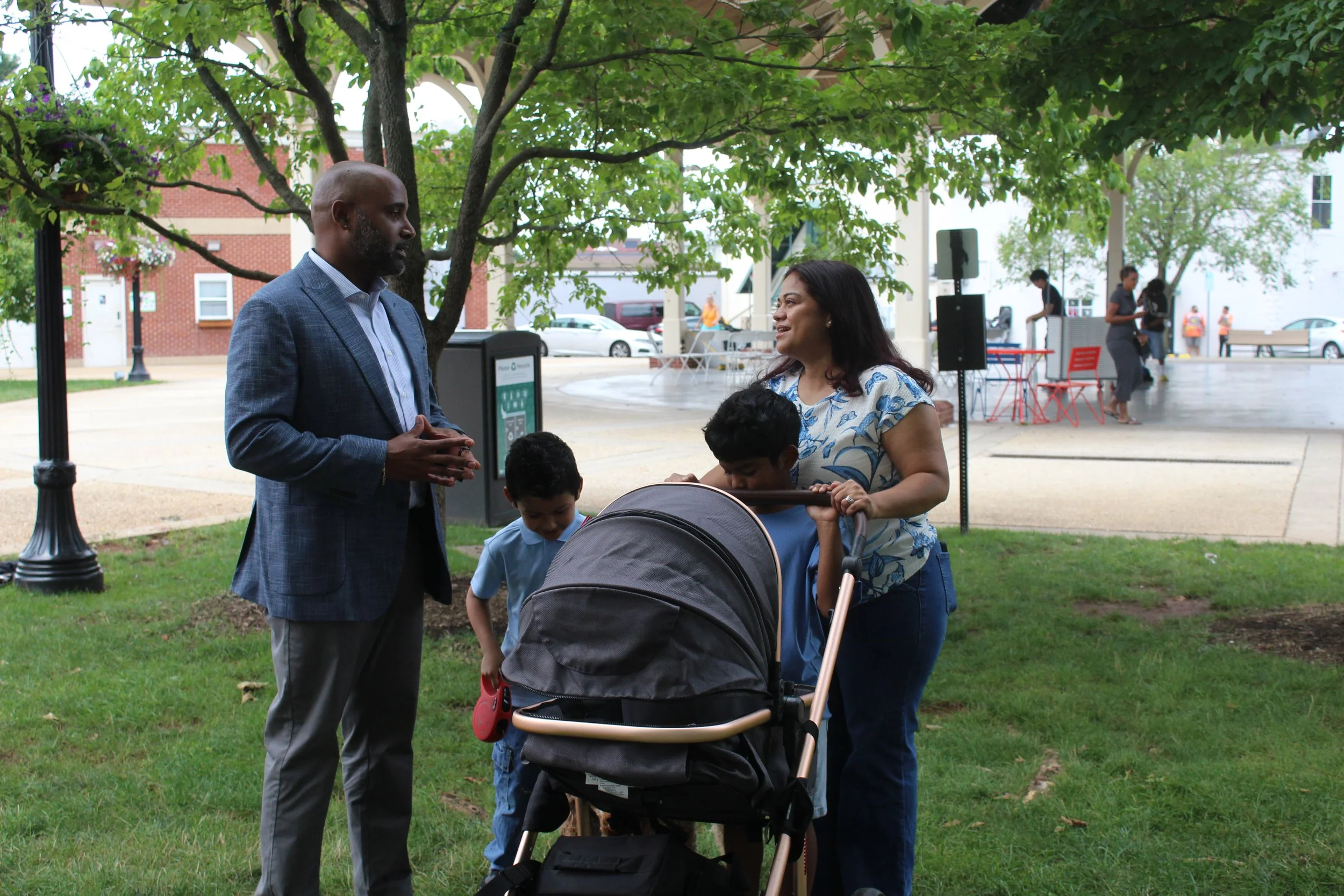 A man in a suit speaking to a woman with a stroller at a park during the daytime, with children and people in the background.