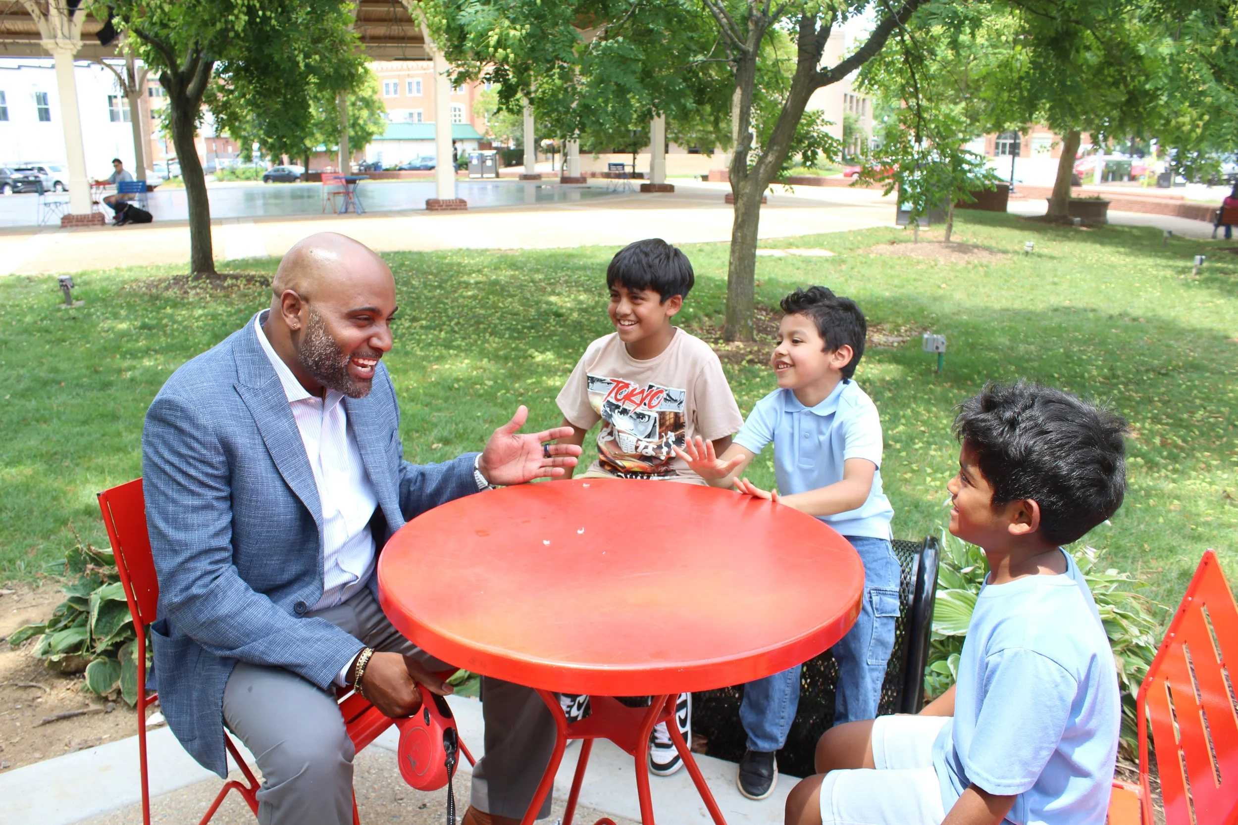 An adult man with a bald head and gray beard wearing a blue blazer and gray pants is sitting at a red outdoor table, talking and gesturing with his hands. Four young boys with dark hair, dressed in casual shirts, are sitting and standing around him, 