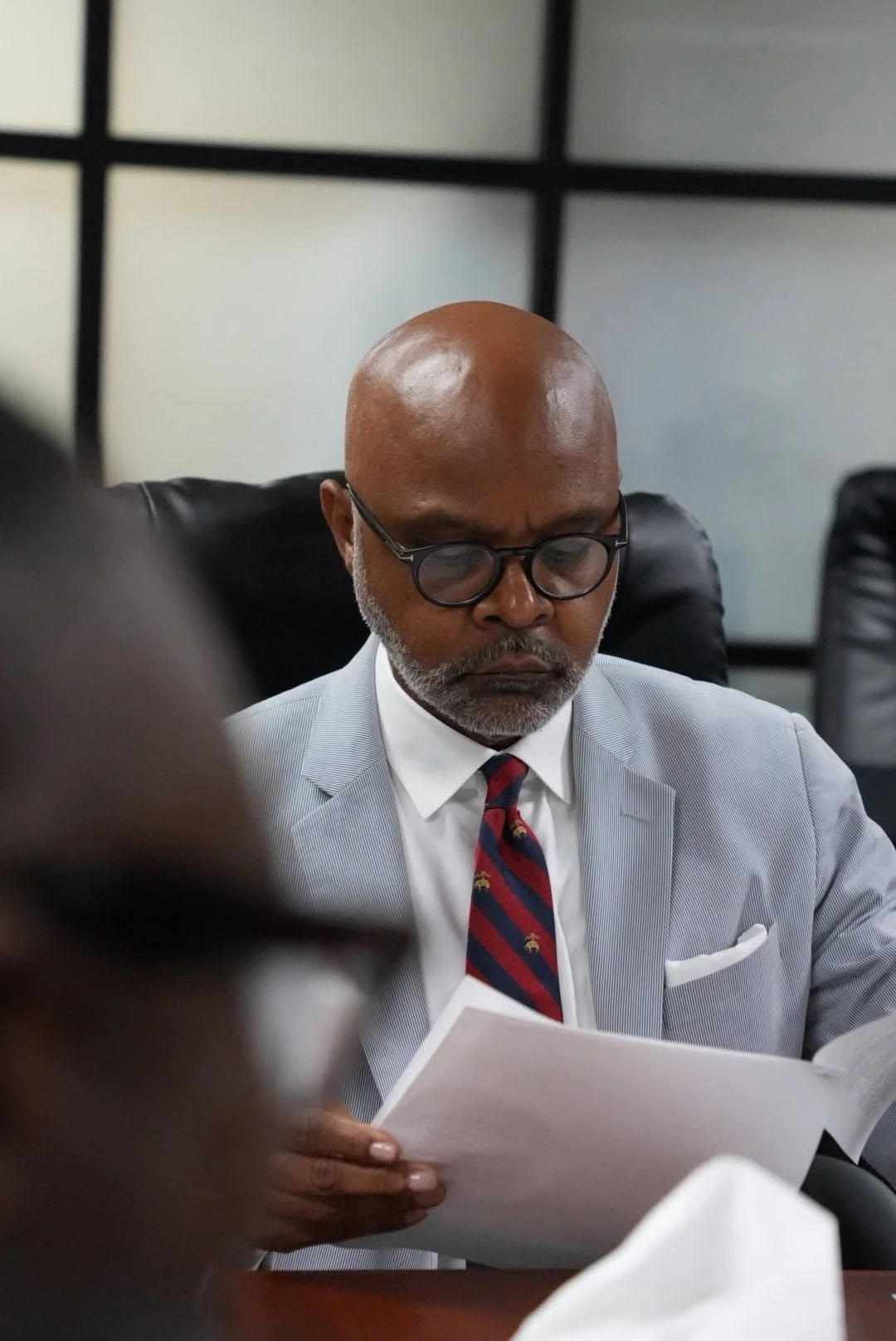 An older African American man with a bald head, glasses, and a gray beard, dressed in a light gray suit with a white shirt and striped red and black tie, reading a document in a modern office setting.