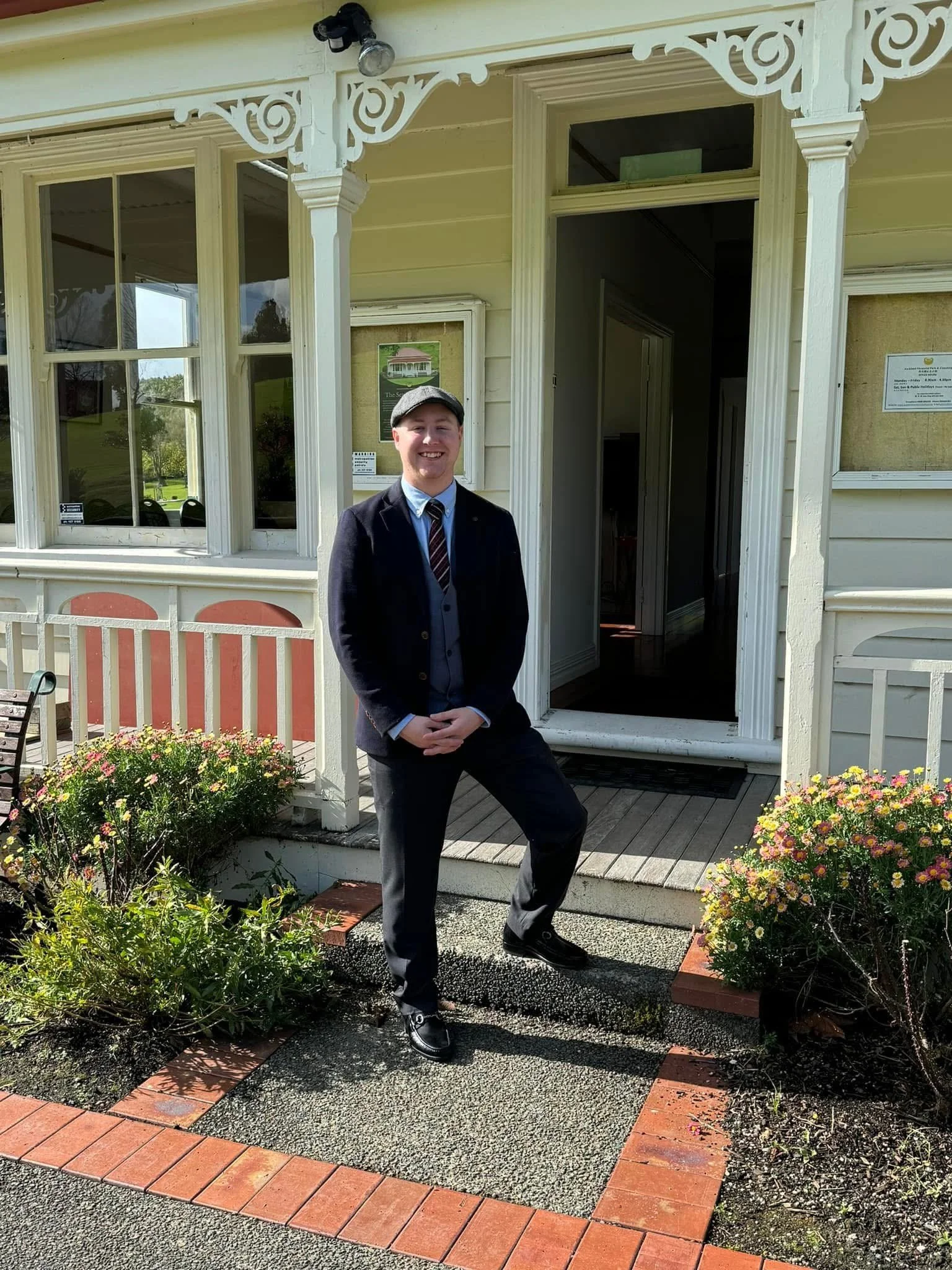 A young man in formal attire, including a suit, tie, and cap, standing in front of a house with decorative white trim and a porch. He is smiling and posing with one leg slightly bent.