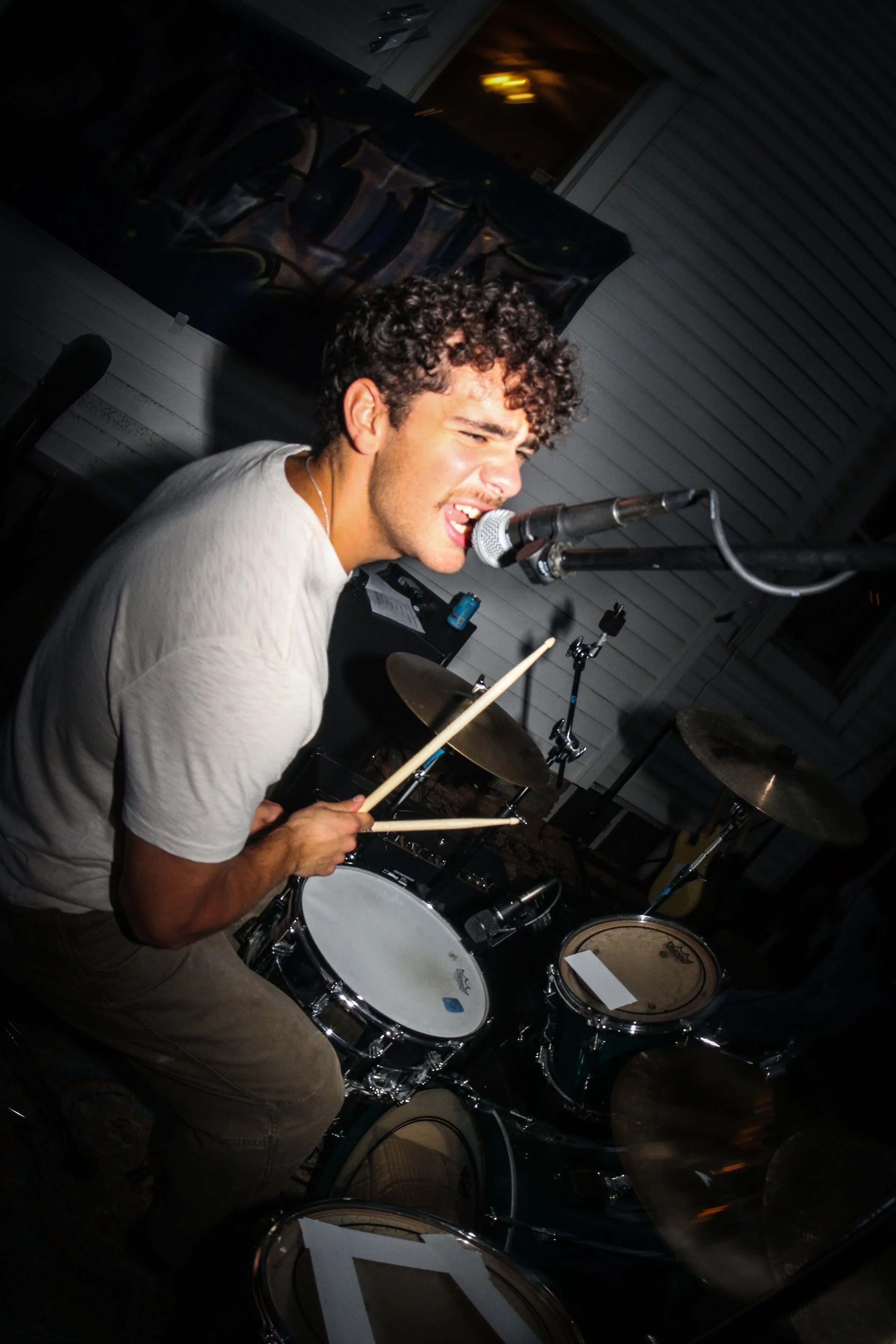 A young man with curly hair singing into a microphone while playing the drums in a dimly lit room.