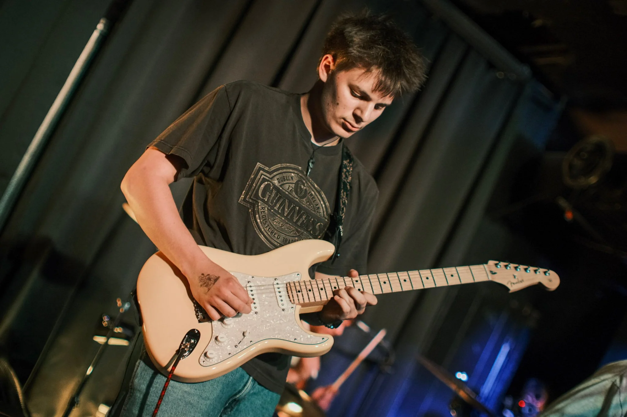 A young man playing an electric guitar on stage, wearing a black T-shirt with a graphic design in a dimly lit venue.