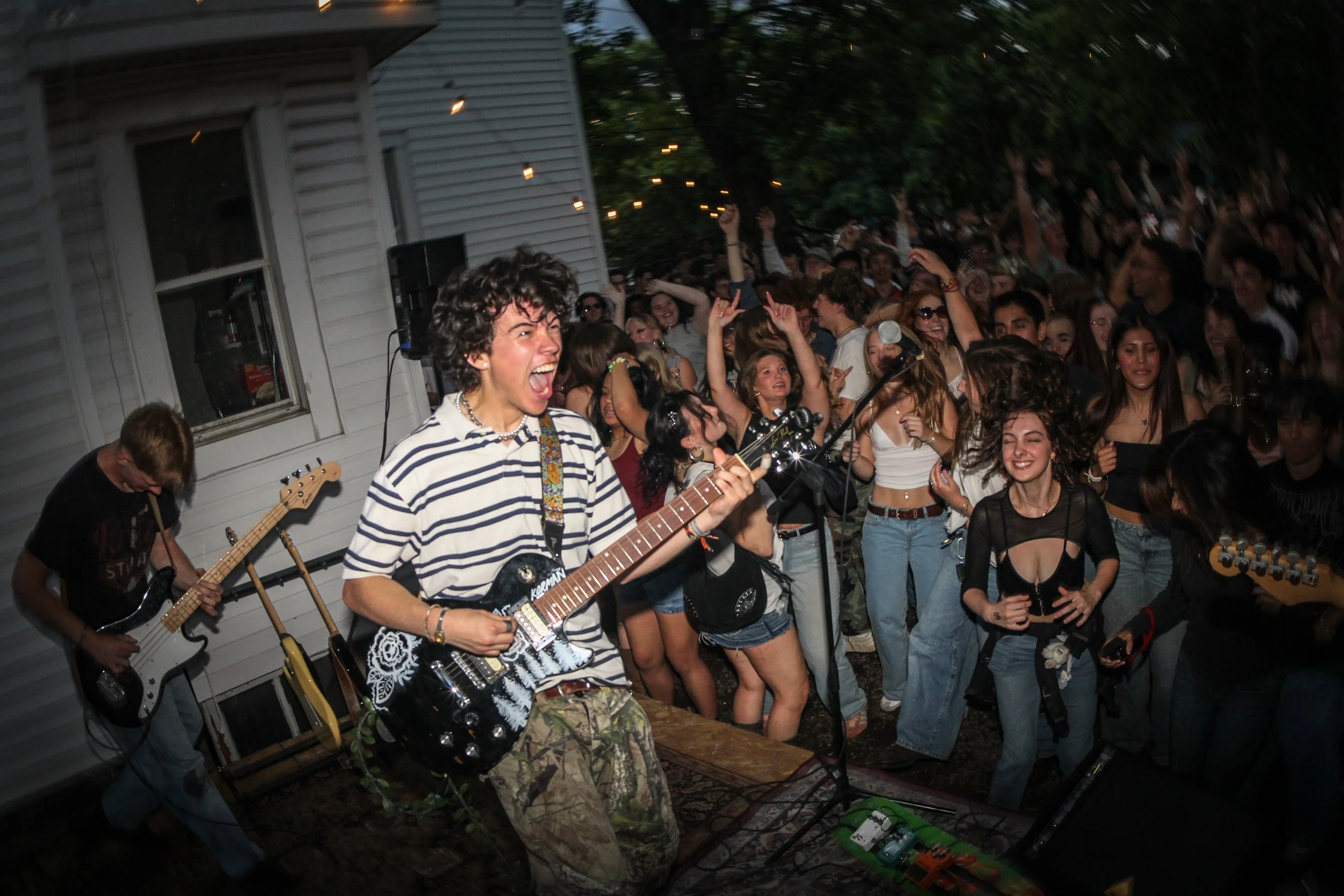 A lively outdoor concert scene with a crowd dancing and having fun, a person playing guitar at the front, and others singing or enjoying the music, set against a background of a house and string lights.