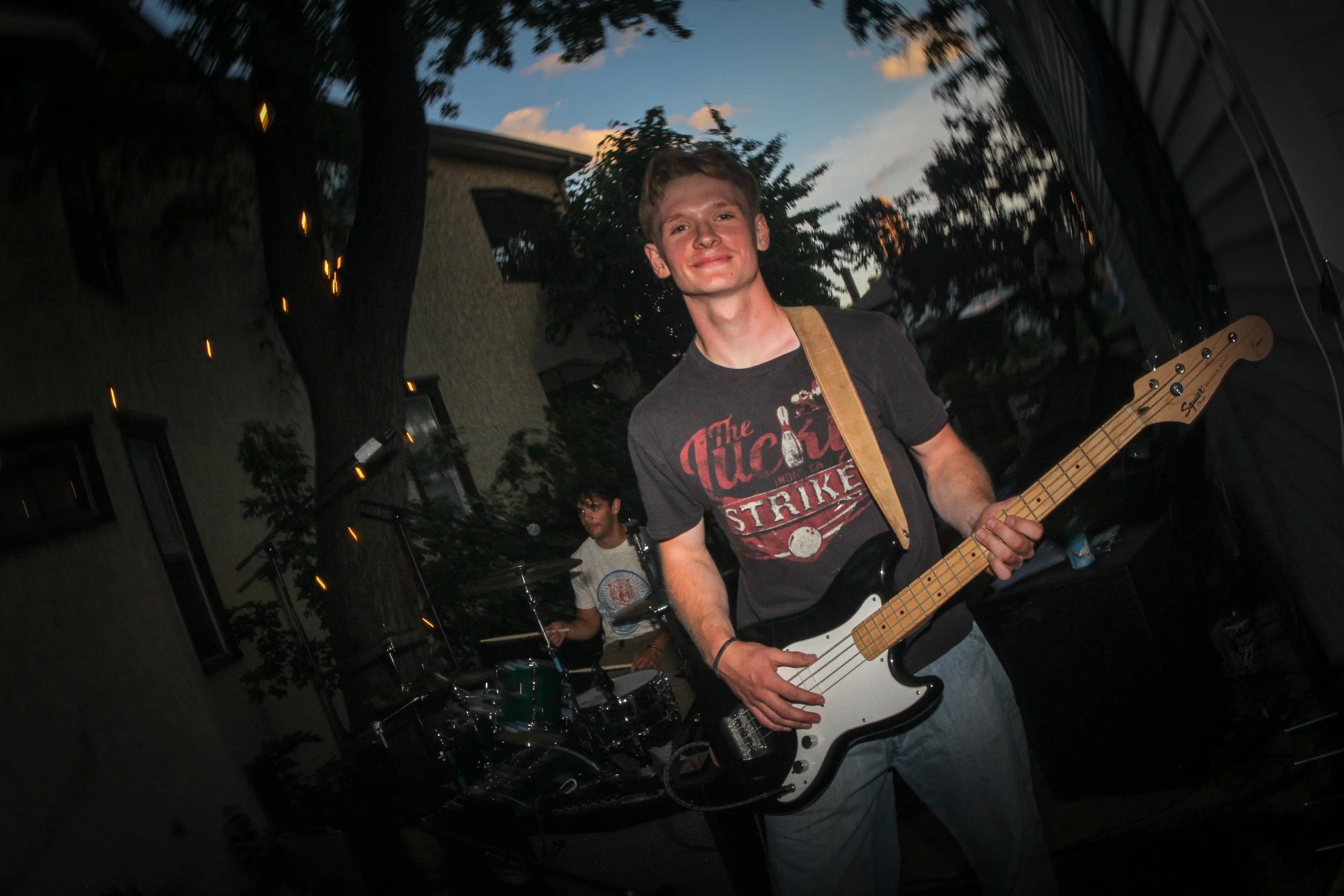 A young man playing an electric bass guitar at an outdoor evening event, with a drummer behind him, under a partly cloudy sky.