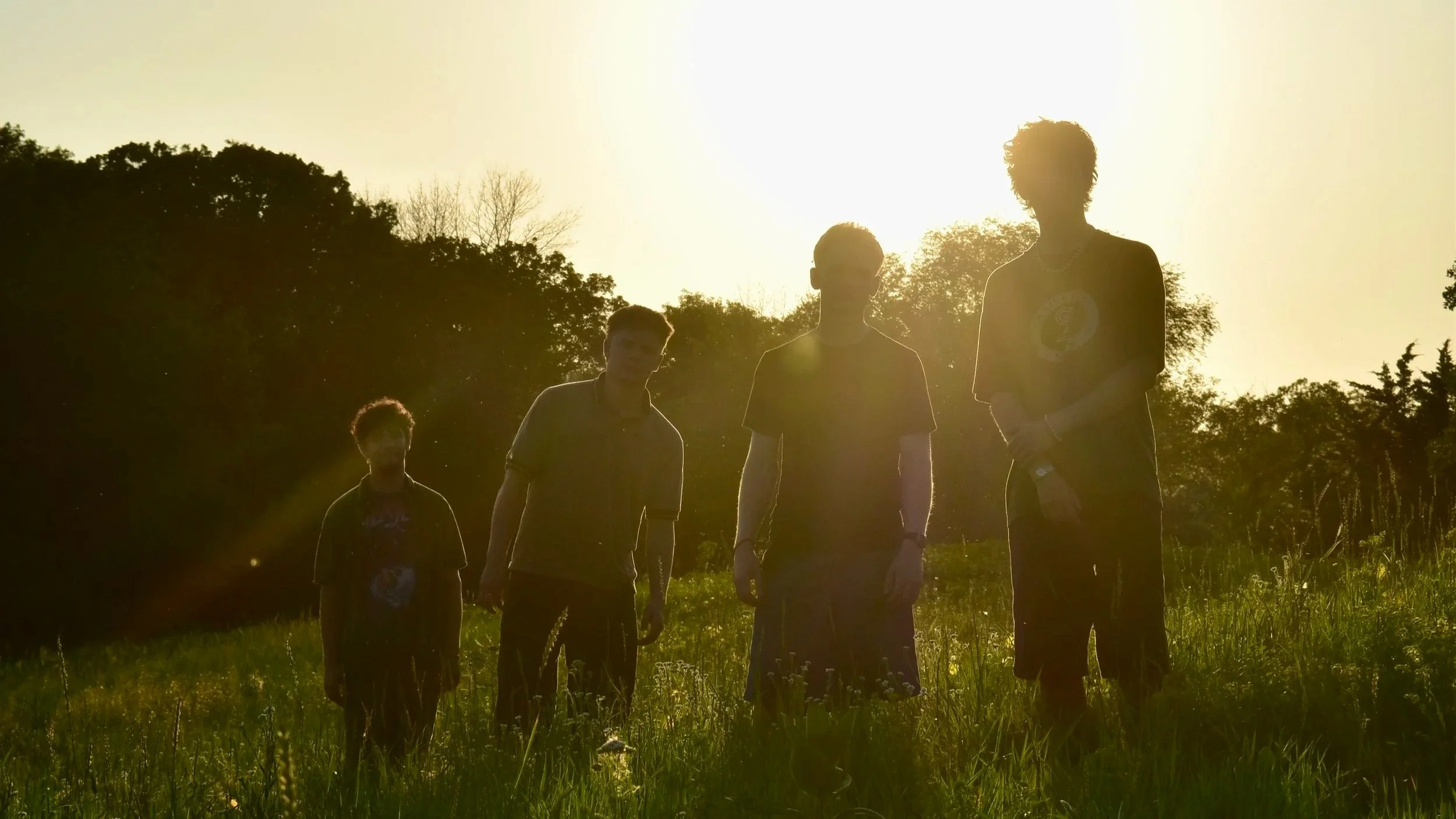 Four people standing outdoors during sunset with silhouettes visible, trees in the background, and a grassy field in foreground.