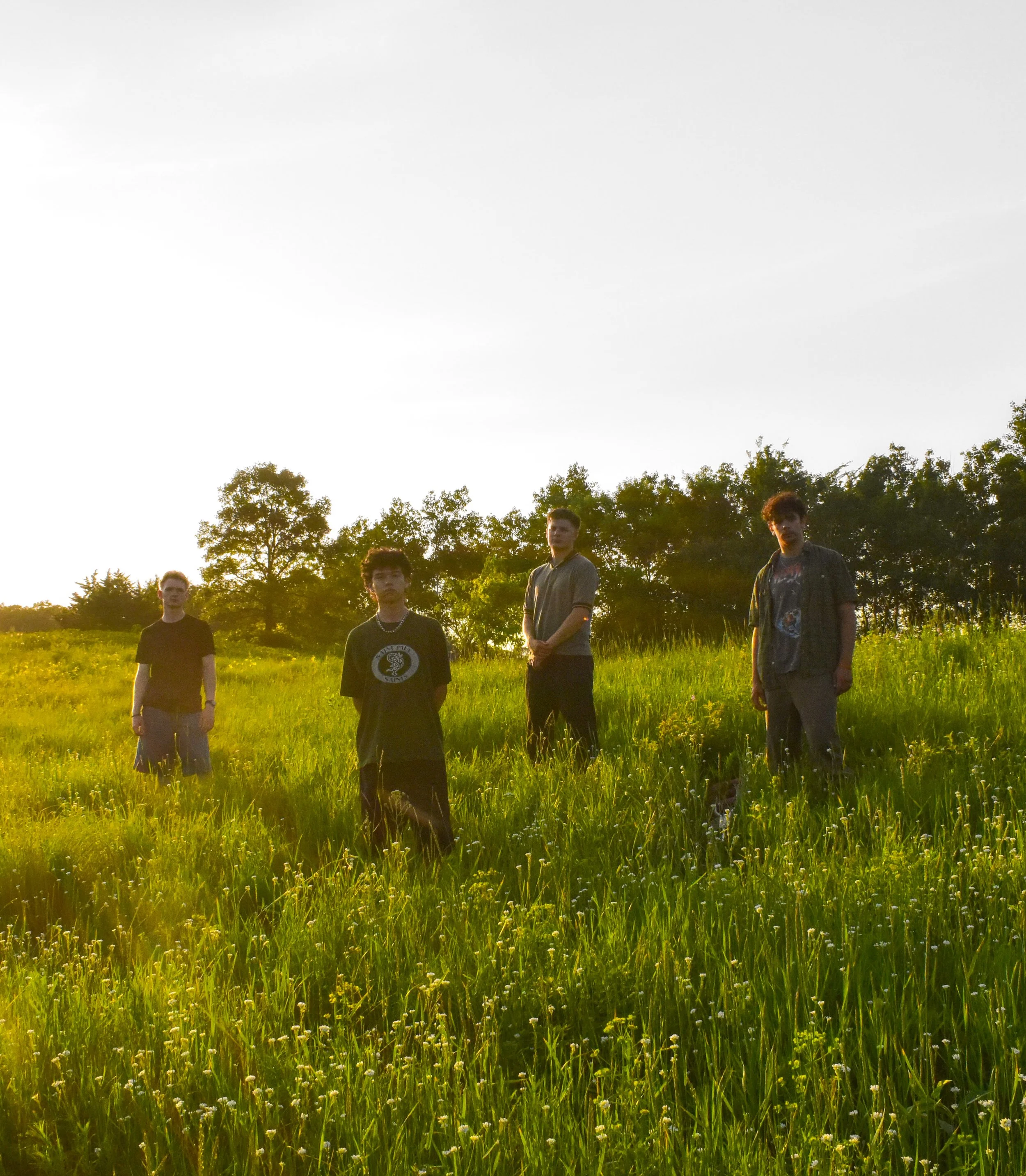 Group of four young guys standing outdoors in a grassy field with trees, during sunset or sunrise.