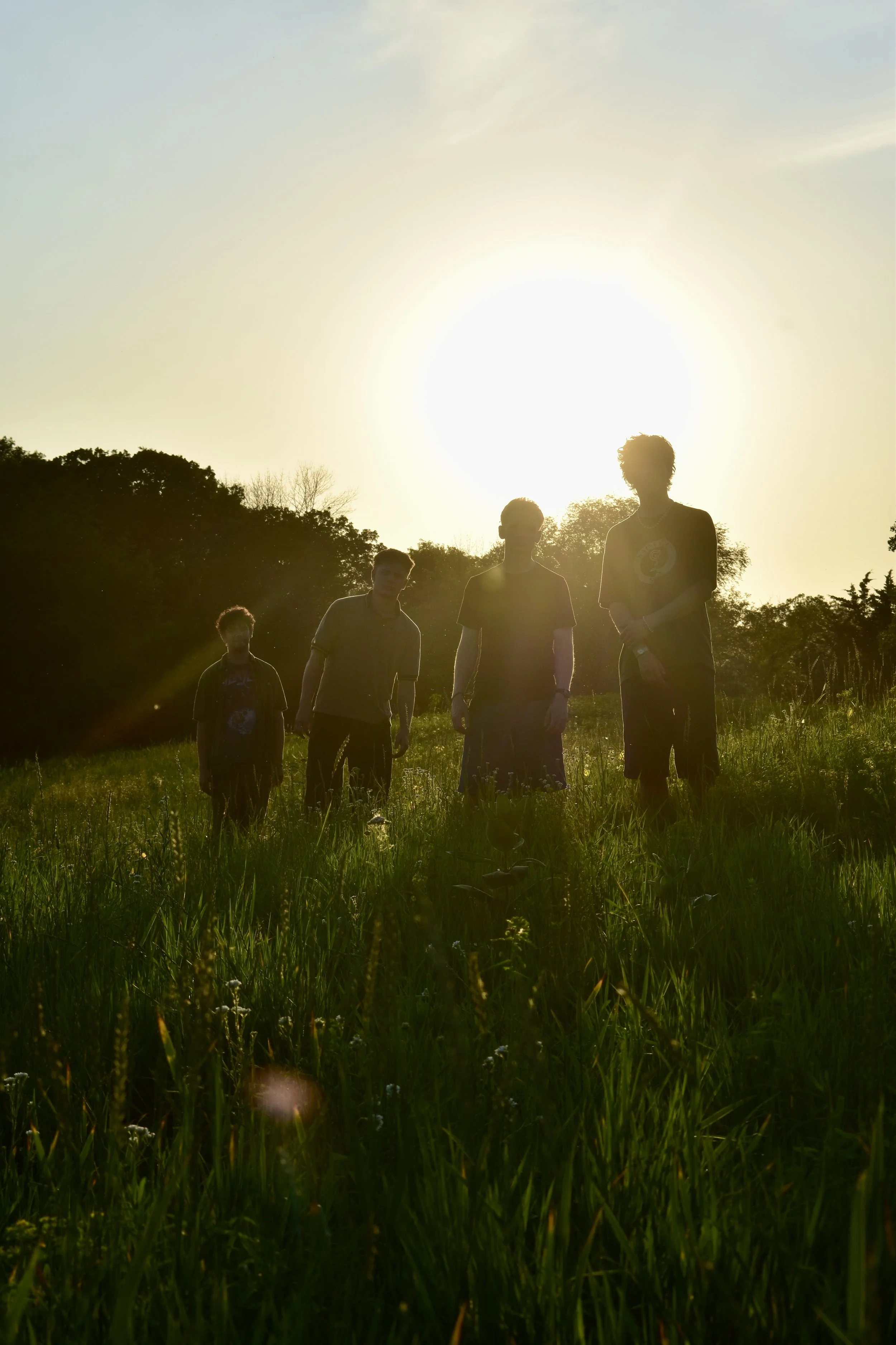 Four young men walking through a grassy field during sunset, with trees in the background and sunlight creating a silhouette effect.
