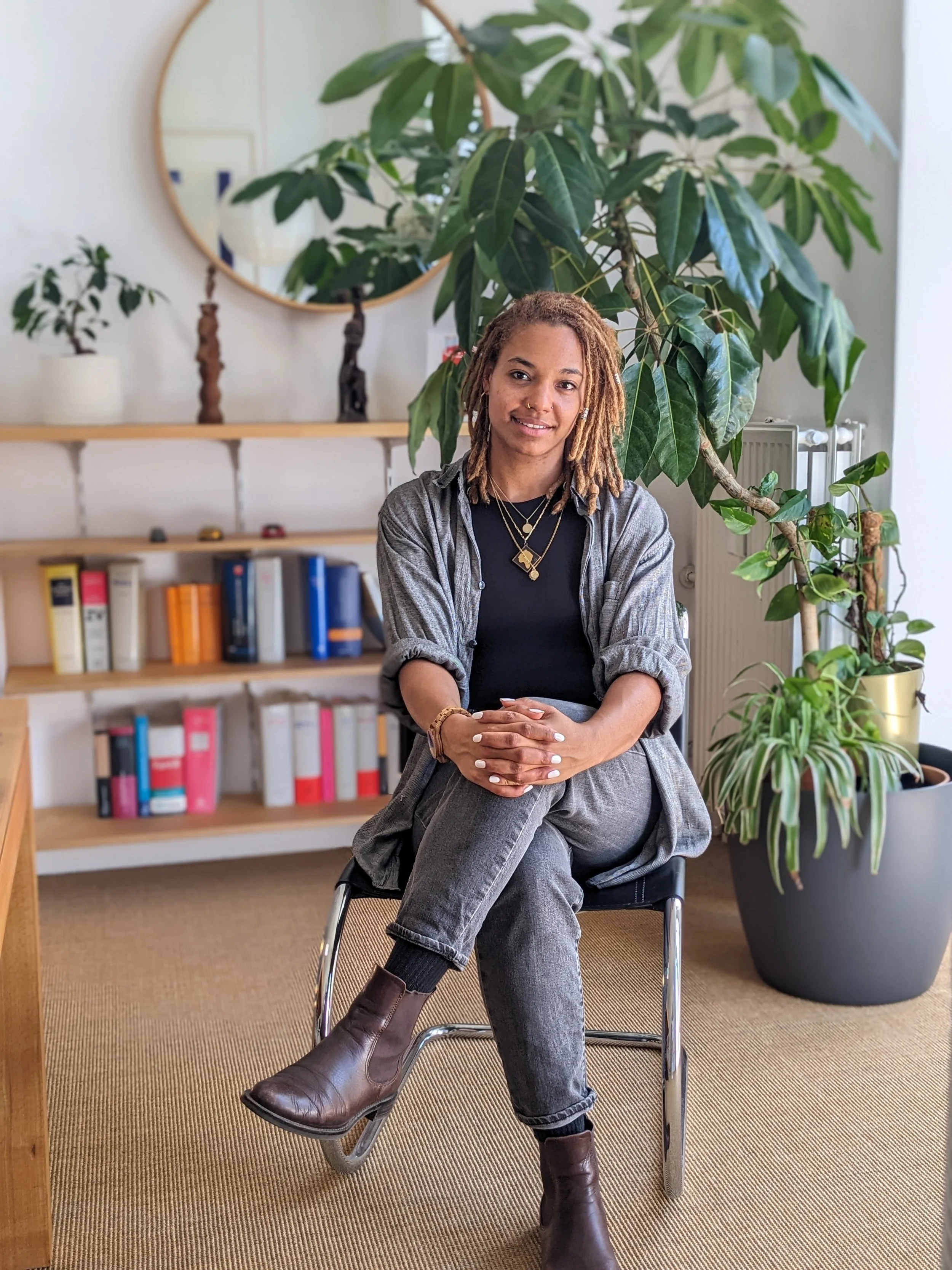 Eine junge Frau mit kurzen Locs sitzt in einem Raum mit Pflanzen und Bücherregalen, trägt einegraue Jacke, schwarze Kleidung, braune Stiefel und mehrere Halsketten, sie lächelt leicht.