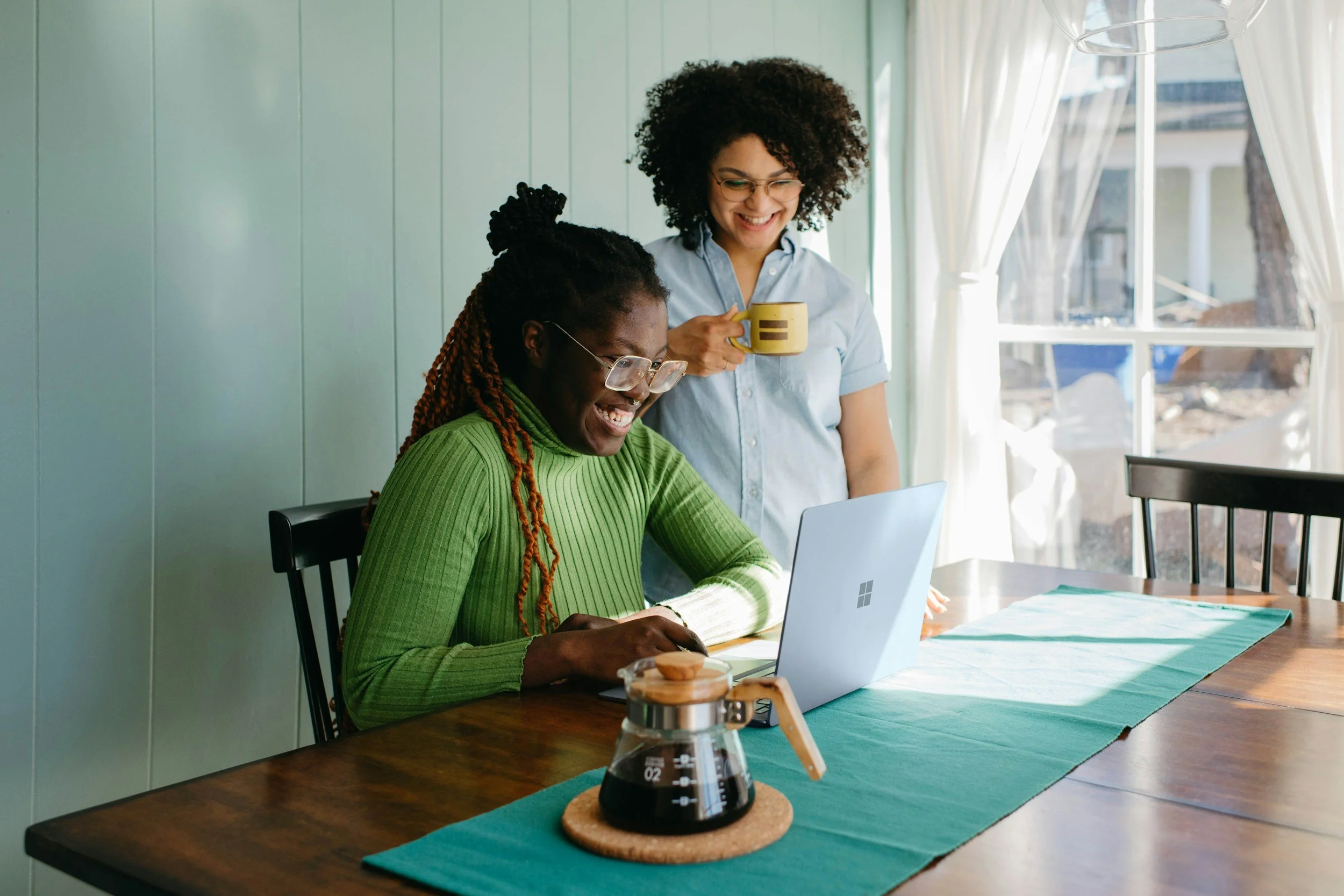 Zwei Frauen arbeiten zusammen an einem Laptop in einem hellen Raum, eine Frau lächelt und hält eine Tasse. Im Vordergrund steht eine Kaffeeampulle auf einem Tisch.