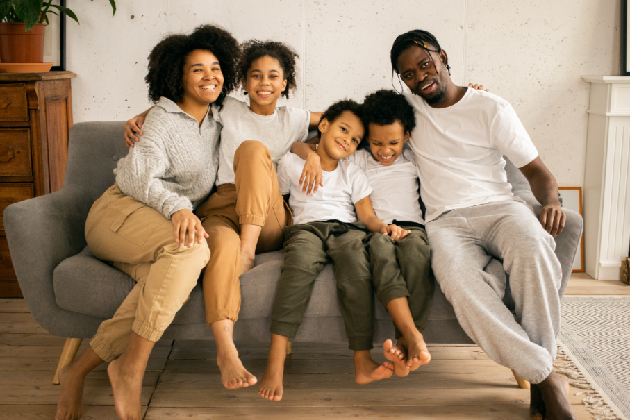 Multiethnische Familie mit zwei Erwachsenen und drei Kindern sitzt lachend auf einem grauen Sofa in einem Wohnzimmer.
