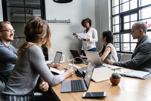Eine Gruppe von fünf Menschen in einem modernen Konferenzraum, einige mit Laptops, die eine Präsentation vor einer Frau halten, die auf einem Whiteboard steht.