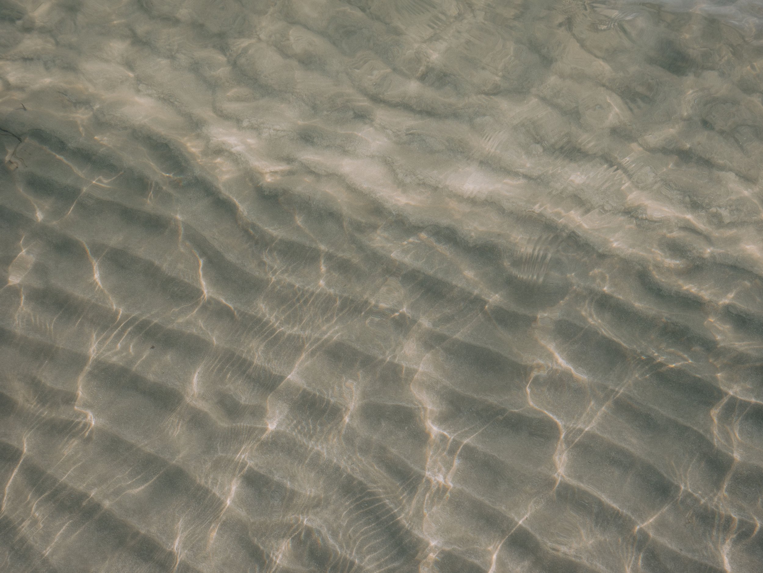 Close-up of water with sunlight reflections creating wavy, shimmering patterns on a sandy or gravelly surface underneath.