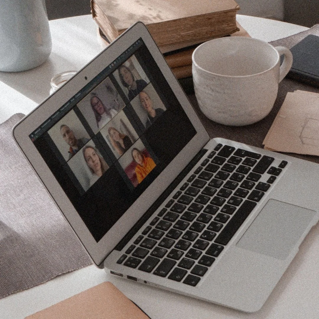 A laptop on a desk showing a video call with nine participants. The desk has a white mug, a large book, some papers, and a cloth placemat.