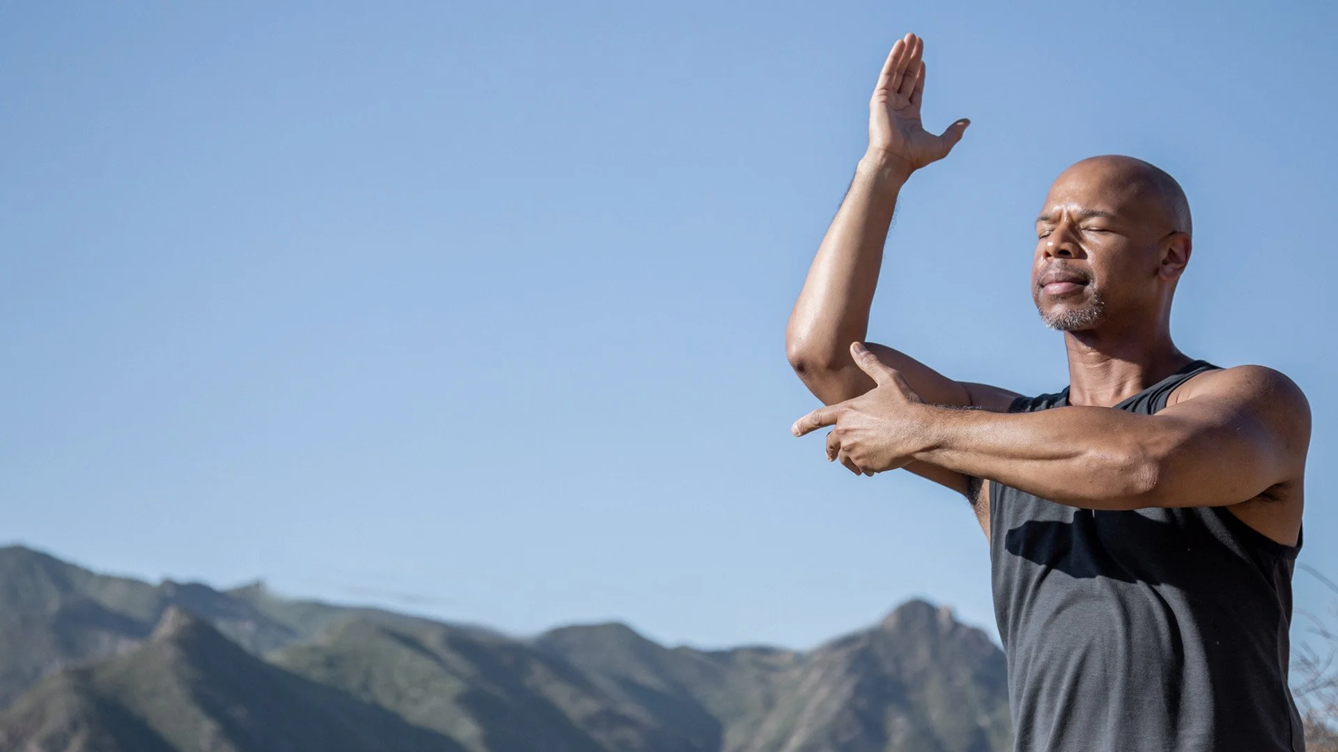 Steven Washington practicing Qigong outdoors with arms crossed, against a mountain backdrop