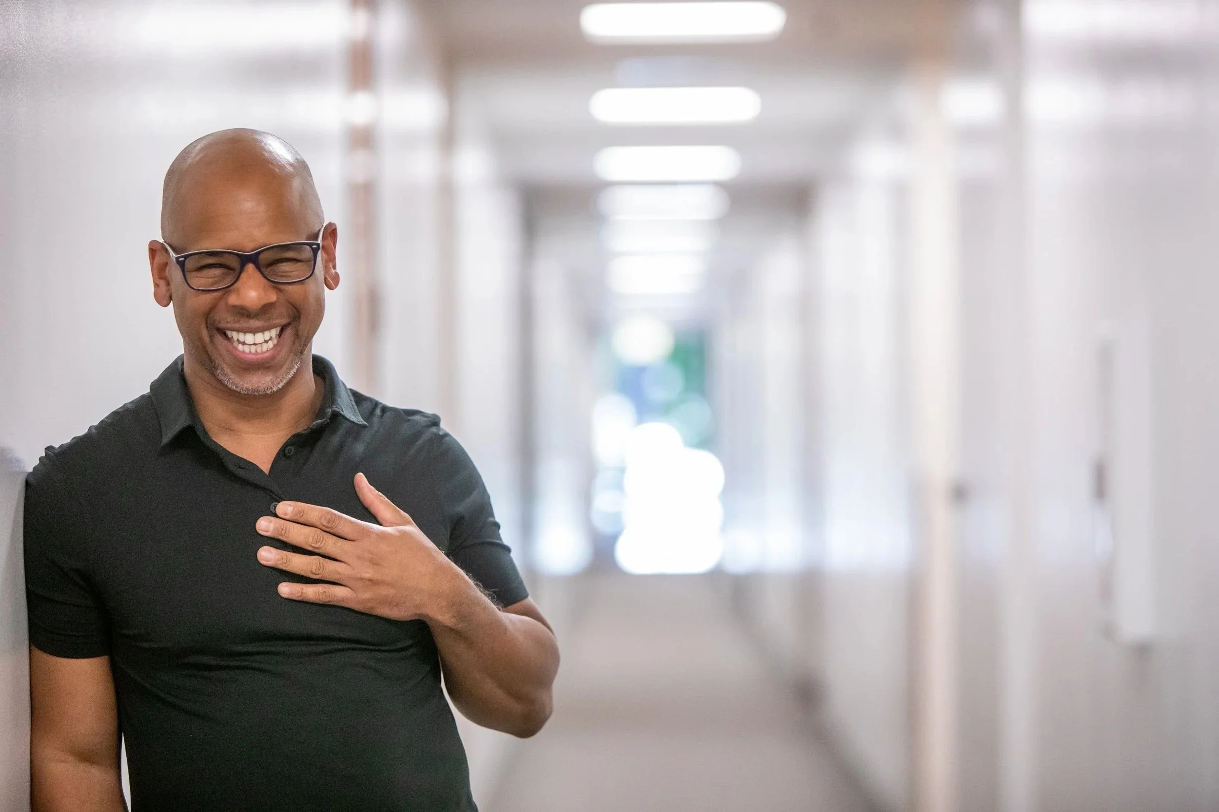 Steven Washington laughing with his hand on his chest in a bright hallway