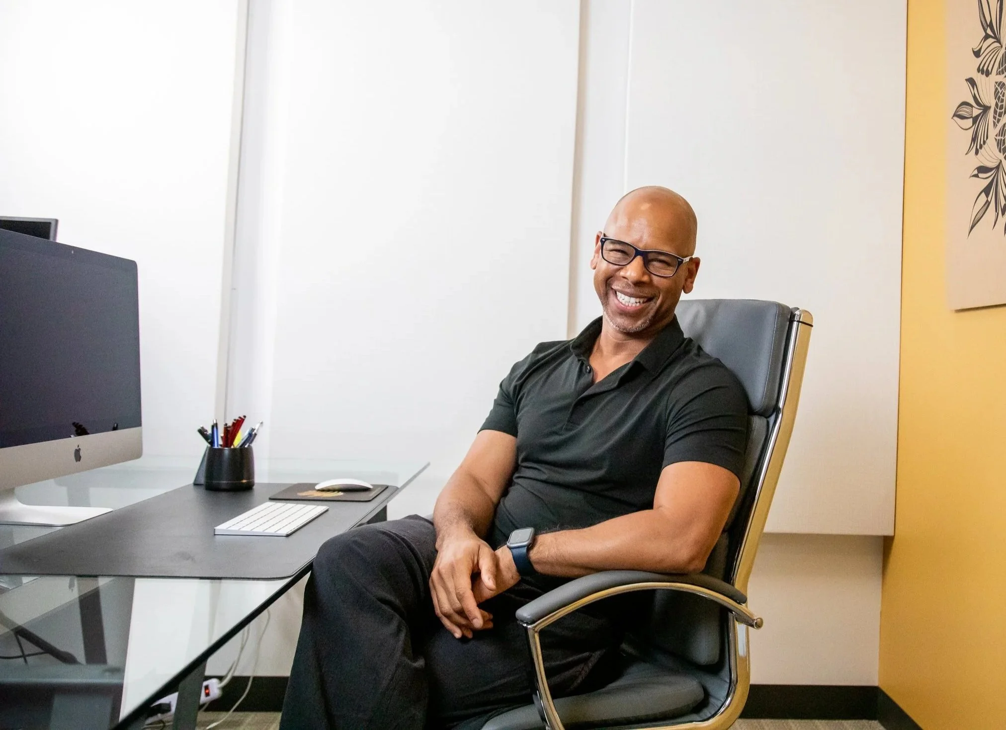 A smiling man with glasses and a smartwatch sitting in an office chair at his desk with a computer, keyboard, mouse, pencils, and a plant pen holder.