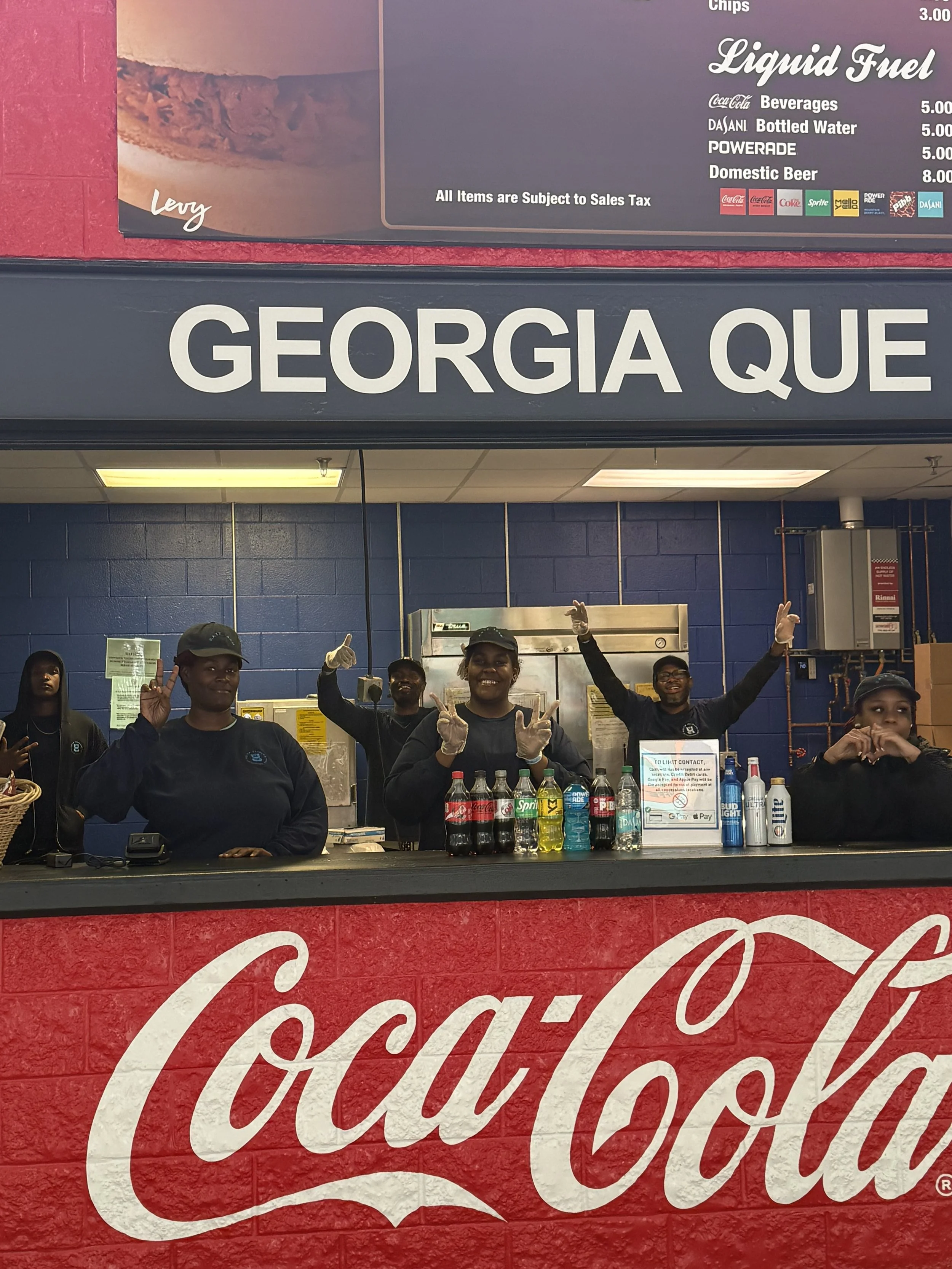 Cash register counter with four people behind it, smiling and making peace signs. Behind them, a blue brick wall and various drink bottles on the counter. Above, a sign displays drink prices and items, with a partial view of a Coca-Cola logo on a red brick wall.