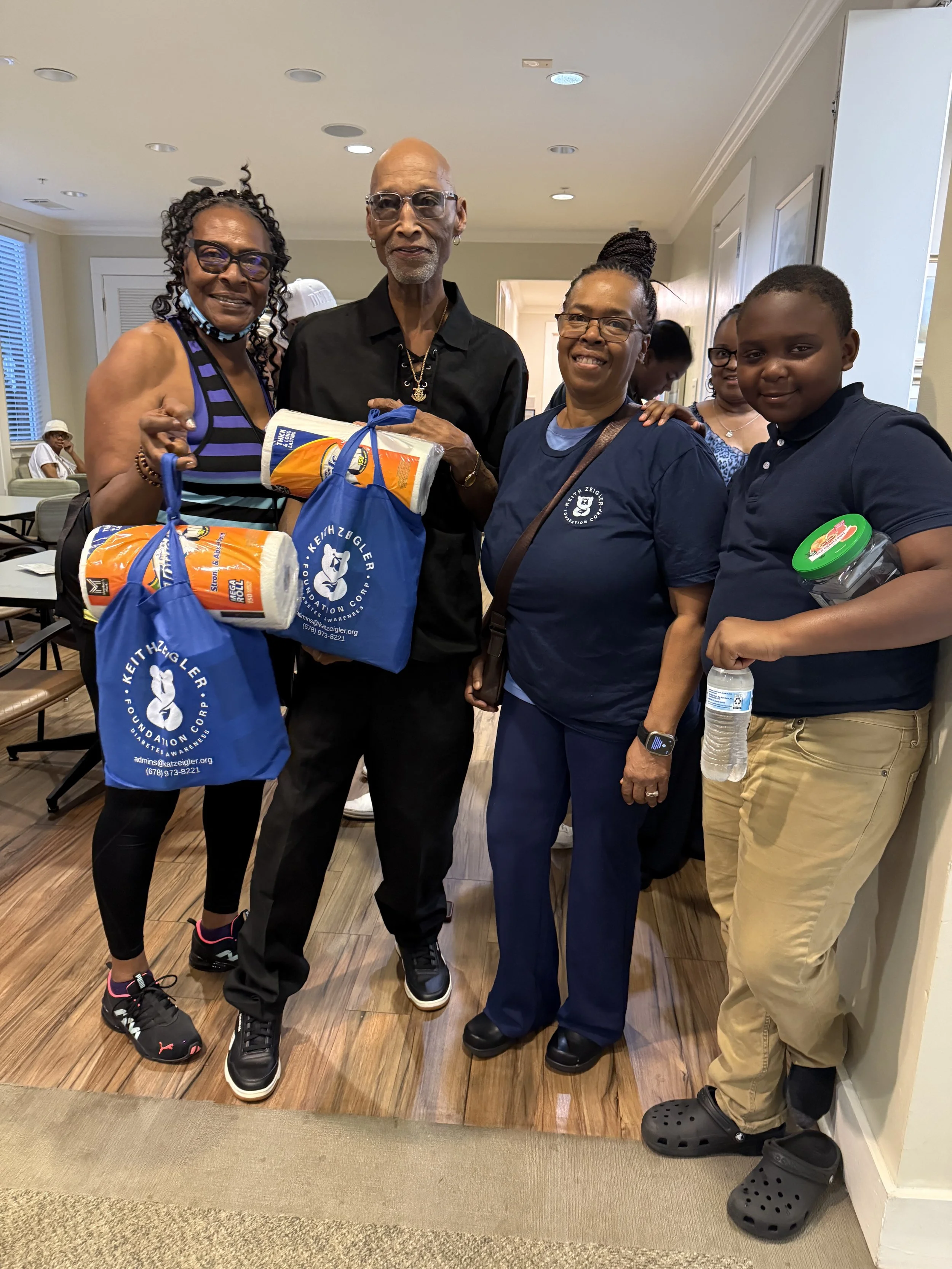A group of five diverse people standing together indoors, holding bags with the Keih Ziegler Foundation logo, smiling at the camera.