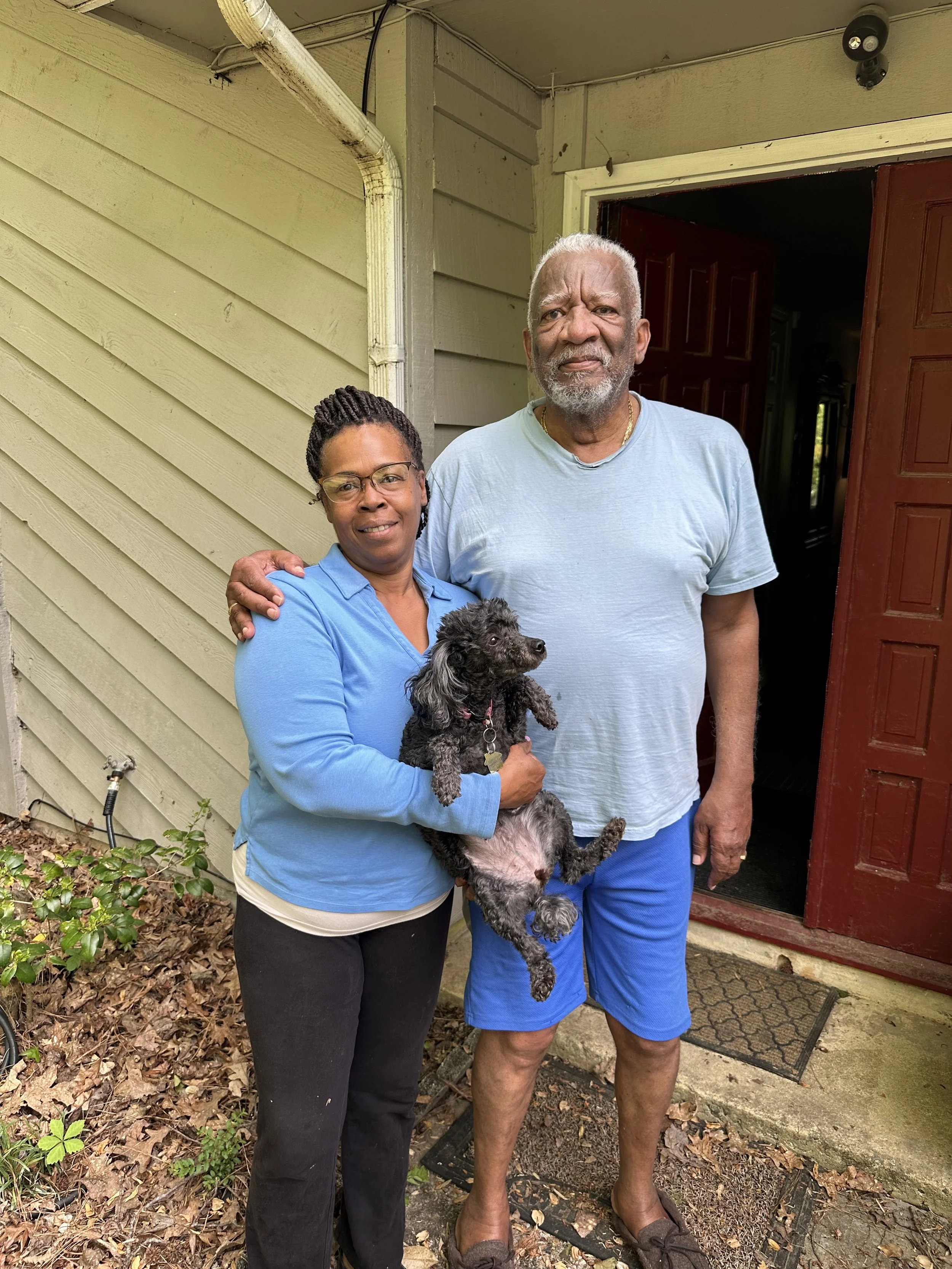 A woman and an older man standing together outside a house, holding a small black and gray dog. The woman is wearing glasses and a blue long-sleeve shirt, and the man is dressed in a light blue t-shirt and blue shorts, with slippers. They are smiling and standing near the entrance of the house, which has a red door and beige siding.
