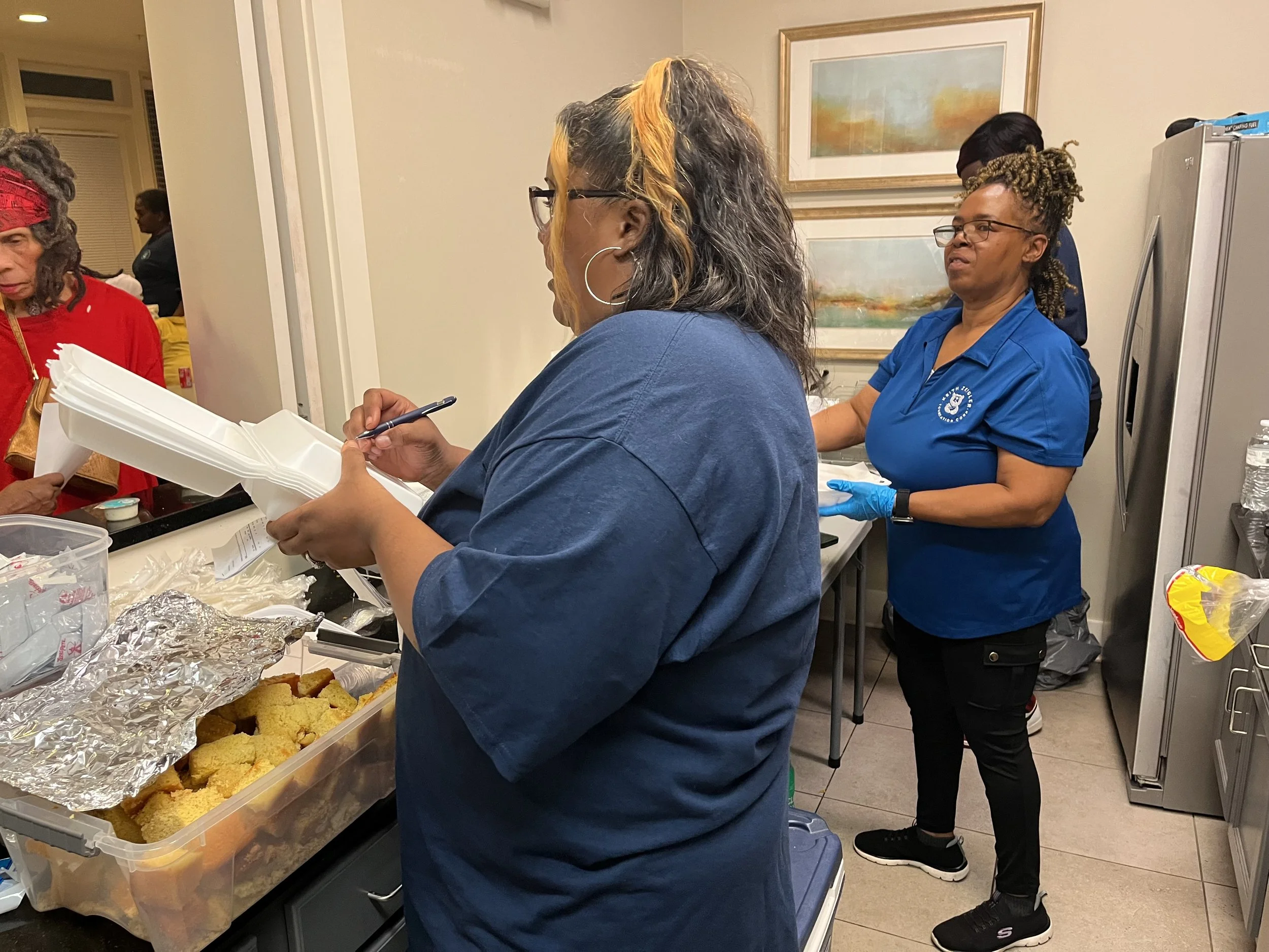 People serving food at a buffet table in an indoor setting, with trays of baked goods and foil-covered dishes, and a woman in a blue shirt and glasses talking to a woman with curly hair with blonde streaks.