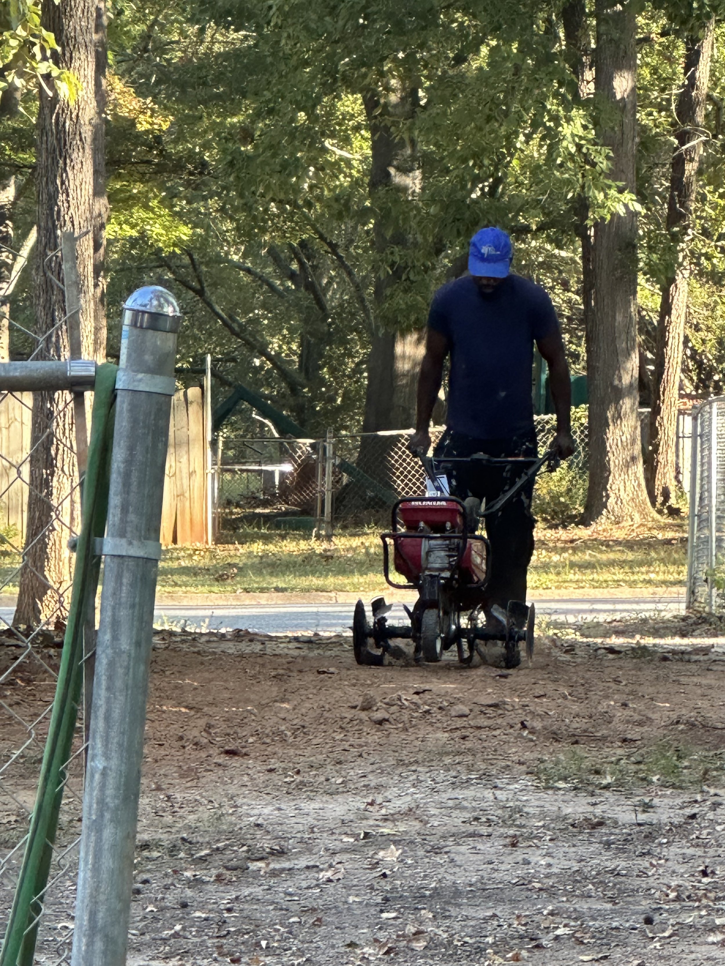 A man mowing a yard with a gas-powered lawn mower near a chain-link fence surrounded by trees.