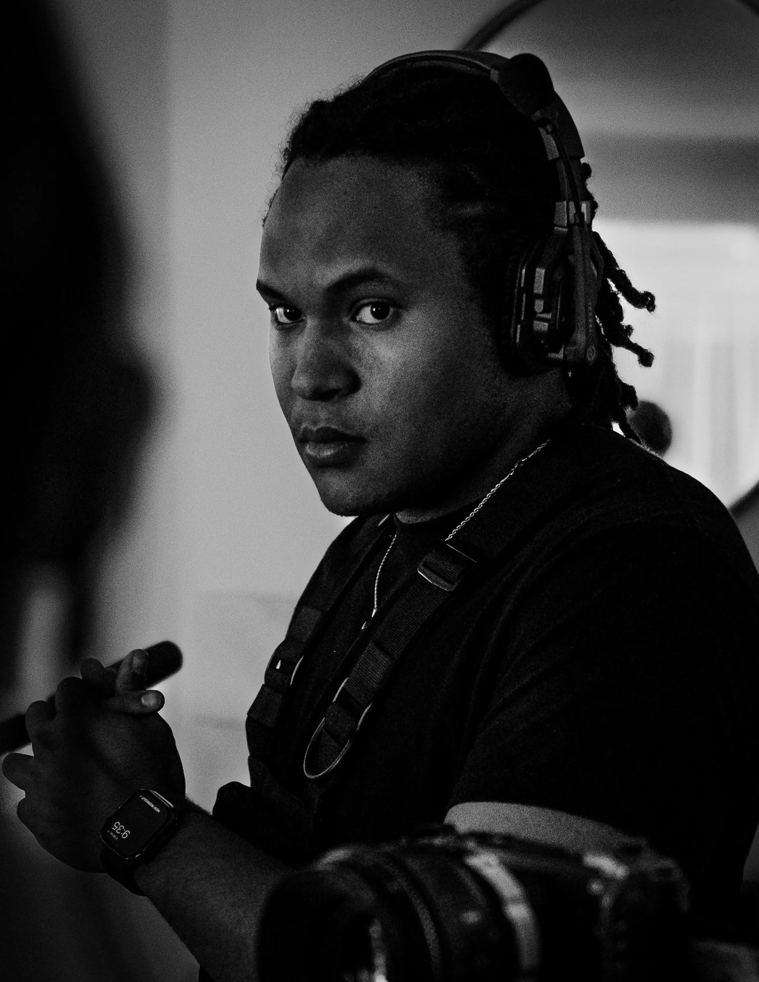 A young man with dreadlocks and a serious expression, wearing headphones, a wristwatch, and carrying a backpack, standing indoors.