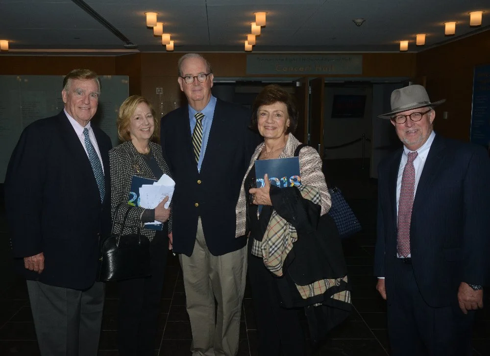 Five people standing together inside a building, dressed in formal attire, smiling at the camera, holding programs and papers, with a sign indicating they are at a concert hall.