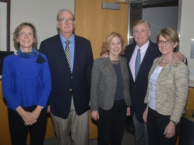 Group of five adults standing indoors, dressed in business attire, smiling for the photo.