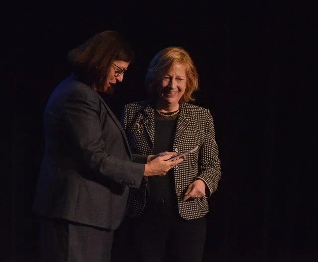 Two women, one with dark hair and glasses and the other with blonde hair, are standing on a dark stage. They are smiling and looking at a smartphone held by the woman with dark hair.