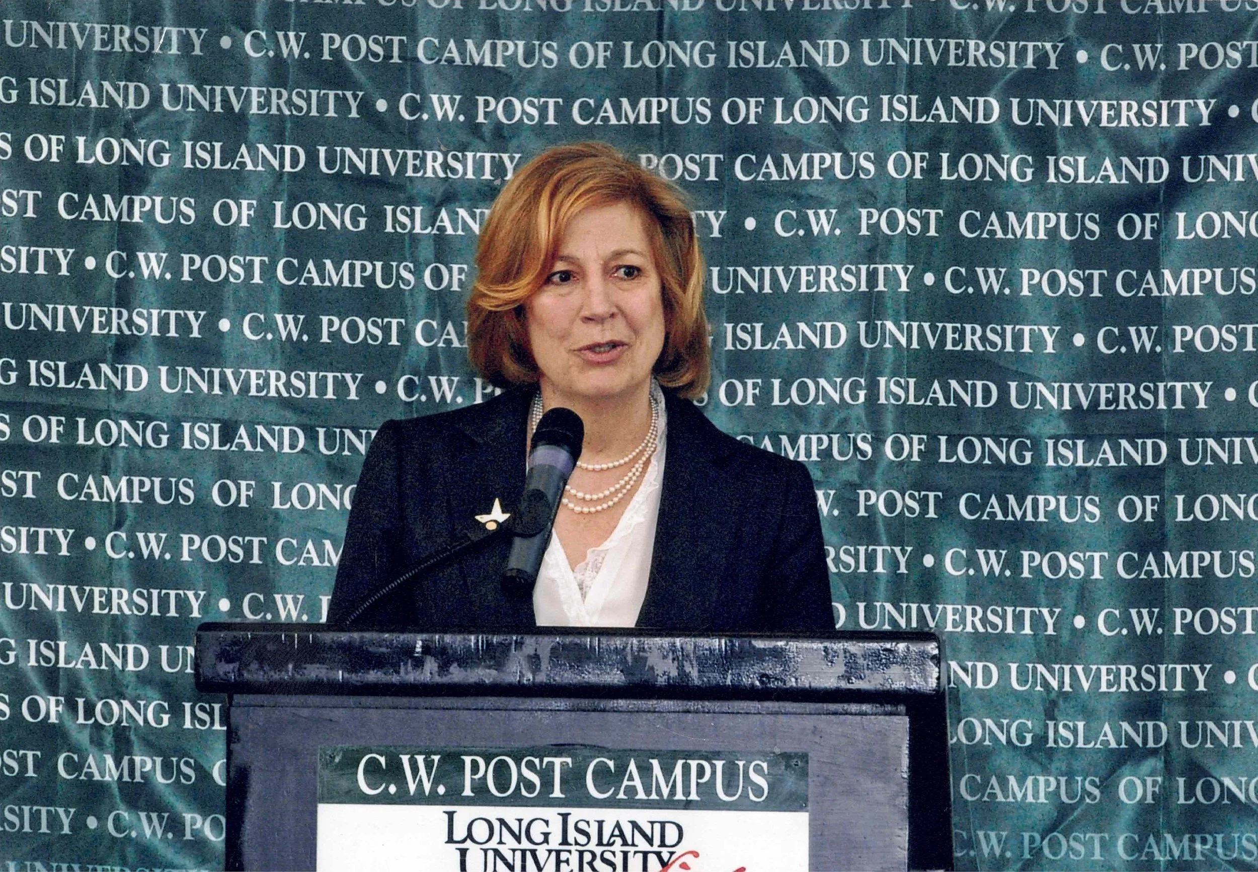 A woman with red hair speaking into a microphone at a podium, standing in front of a blue-green backdrop with white text that reads Post Campus of Long Island University.
