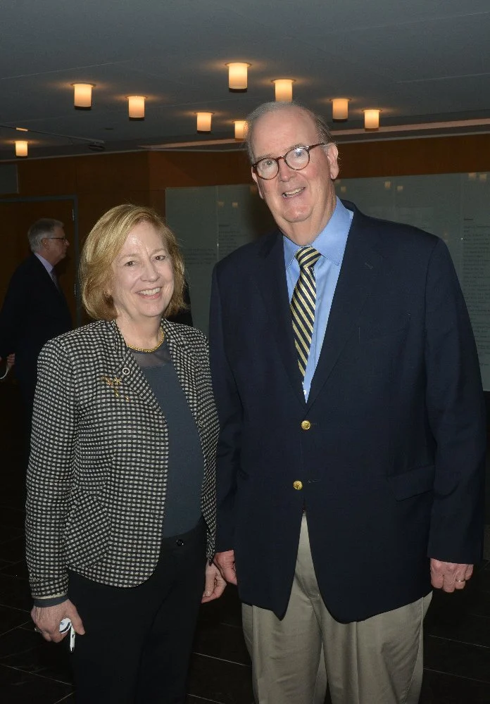 A woman and a tall man in business attire posing for a photo indoors, with other people and informational display boards in the background.
