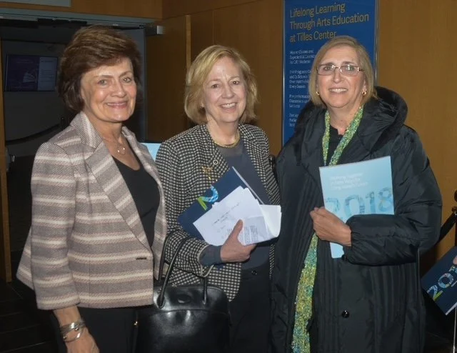 Three women standing together at an event, smiling and holding papers and booklets. A blue informational sign is in the background.