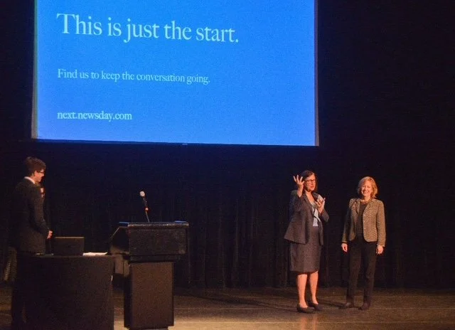 Two women standing on a stage during a presentation or event, with a large screen behind them displaying text that says 'This is just the start.' and a URL 'next.newsday.com'. One woman is speaking into a microphone while the other stands beside her,