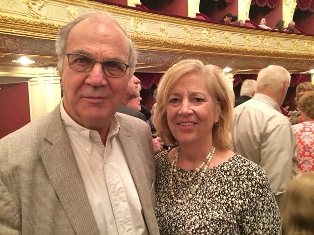 Kostas and Nancy Douzinas posing together in a theater with gold ornate decor and red velvet curtains, surrounded by other people in formal attire.