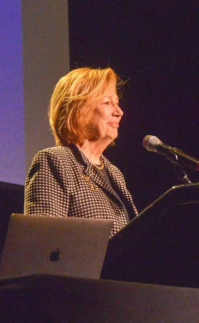 A woman with blonde hair standing at a podium, speaking into a microphone, with a laptop in front of her.