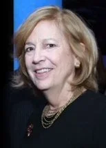 Close-up of a smiling woman with blonde hair, wearing a dark top and gold necklace, against a neutral background.