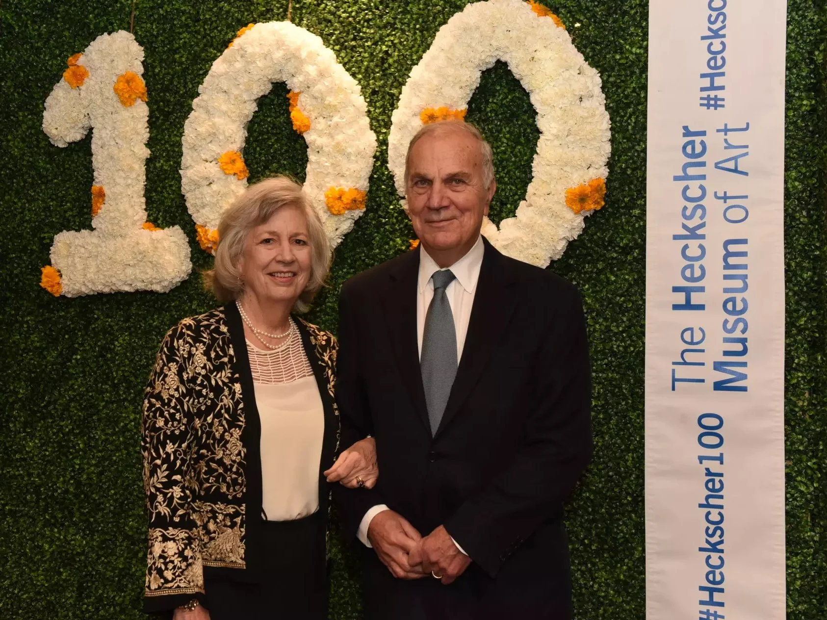 Kostas and Nancy Douzinas standing together at a celebration for the 100th anniversary of the Heckscher Museum, with a green hedge wall and large white flowers spelling '100' behind them. 