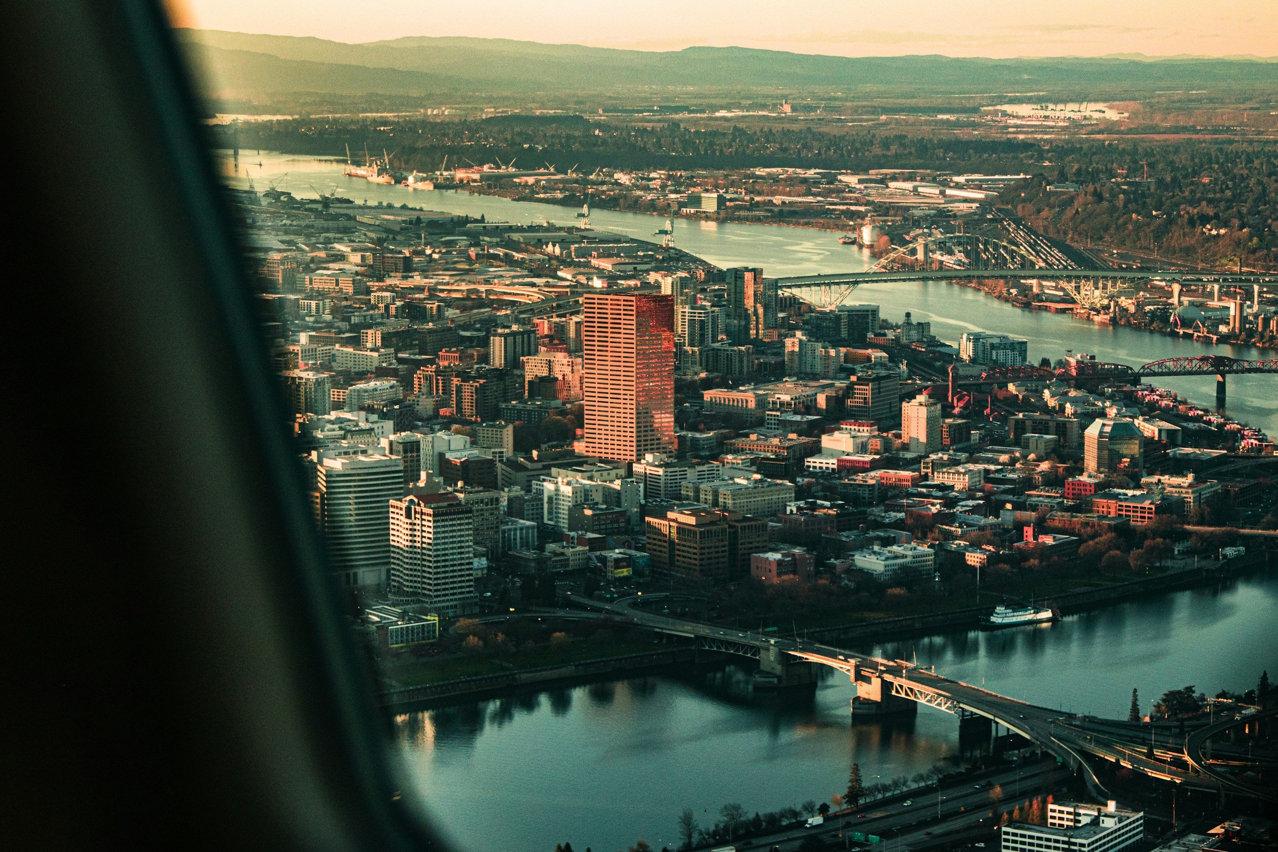 Aerial view of a city with numerous buildings, bridges over a river, and industrial areas in the background, taken from an airplane window during sunset.