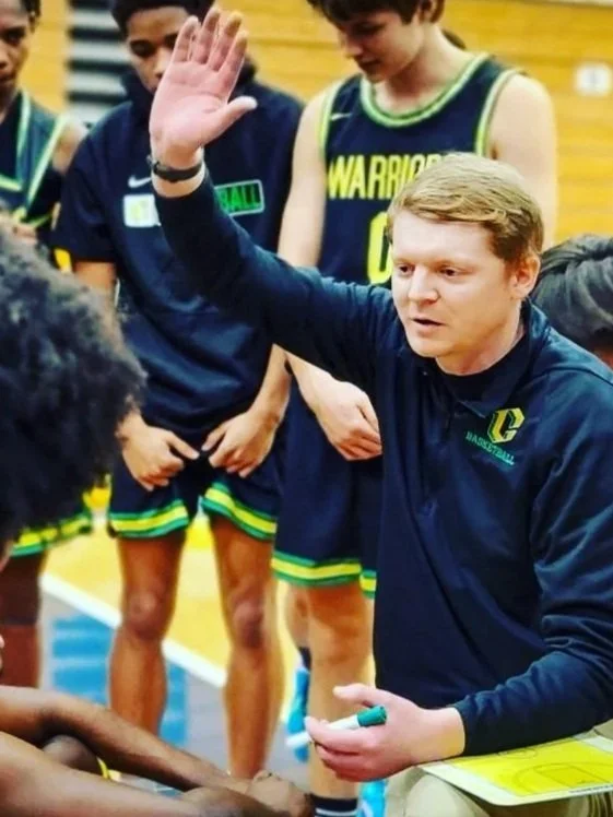 A basketball coach in a navy jacket with a logo is giving instructions to a team of players during a timeout. The players are in dark blue jerseys with green and yellow accents, in a gymnasium.