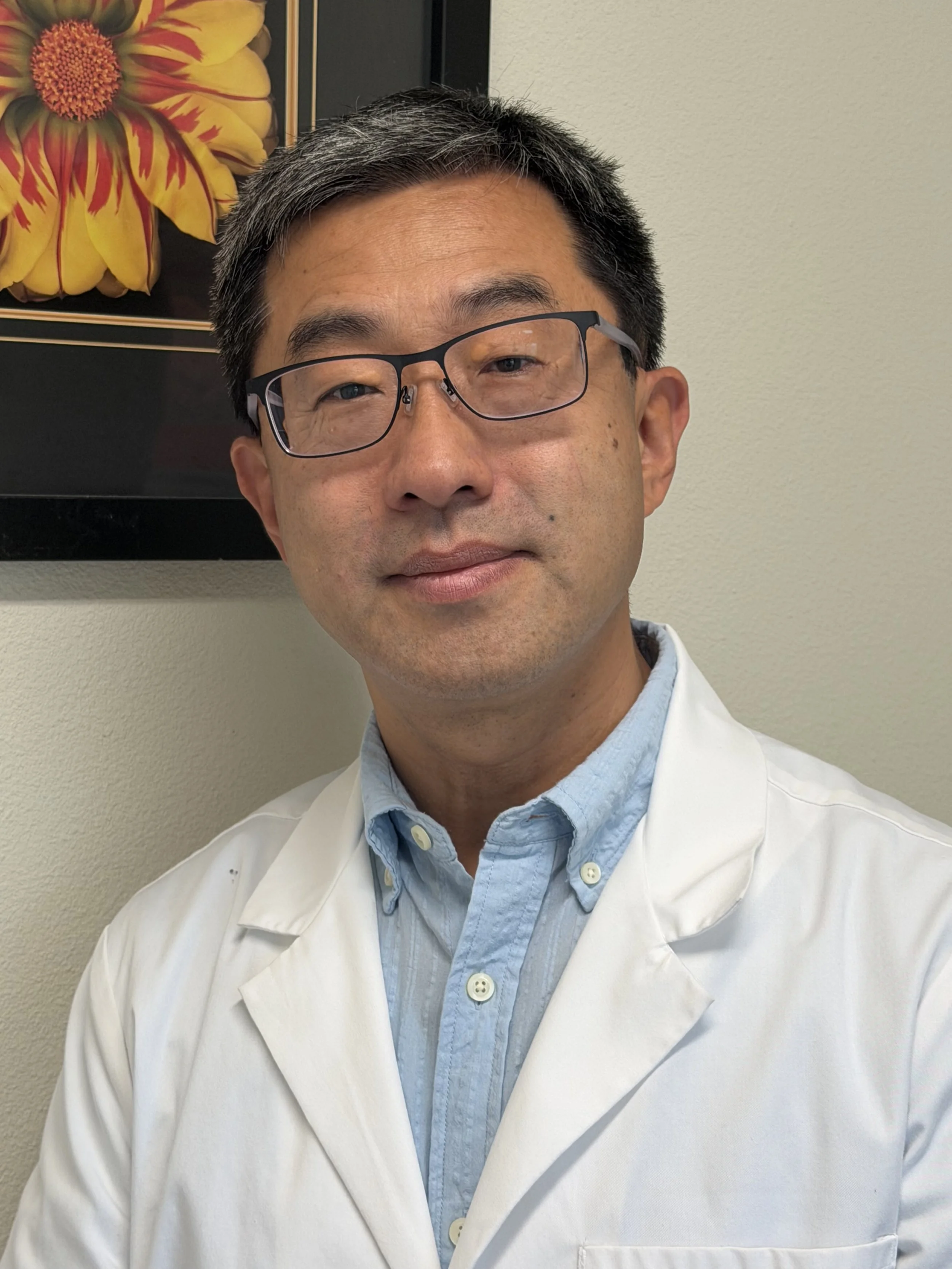 Headshot of an Asian male healthcare professional wearing glasses and a white coat, standing against a plain wall with a framed floral artwork in the background.