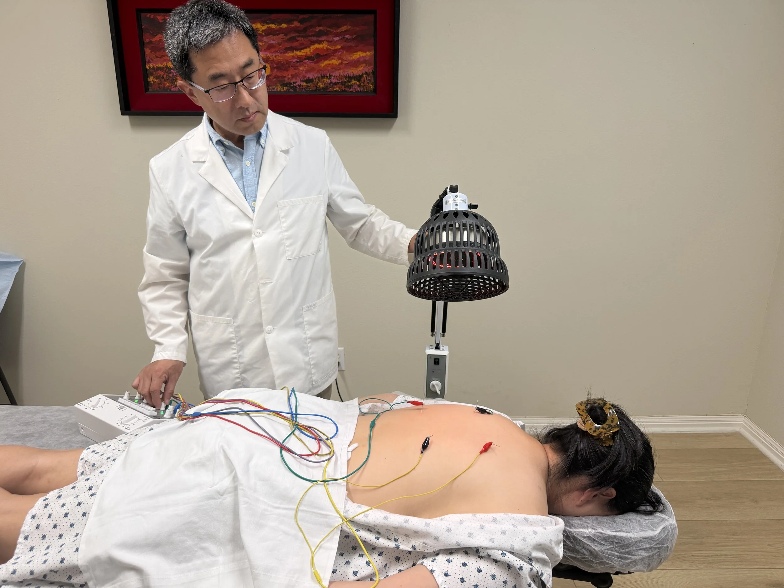 A medical professional performing a procedure on a patient lying face down on a table, with wires attached to the patient's back and a device emitting light below a large lamp. The professional is wearing glasses and a white lab coat and is standing beside the patient in a clinical setting.
