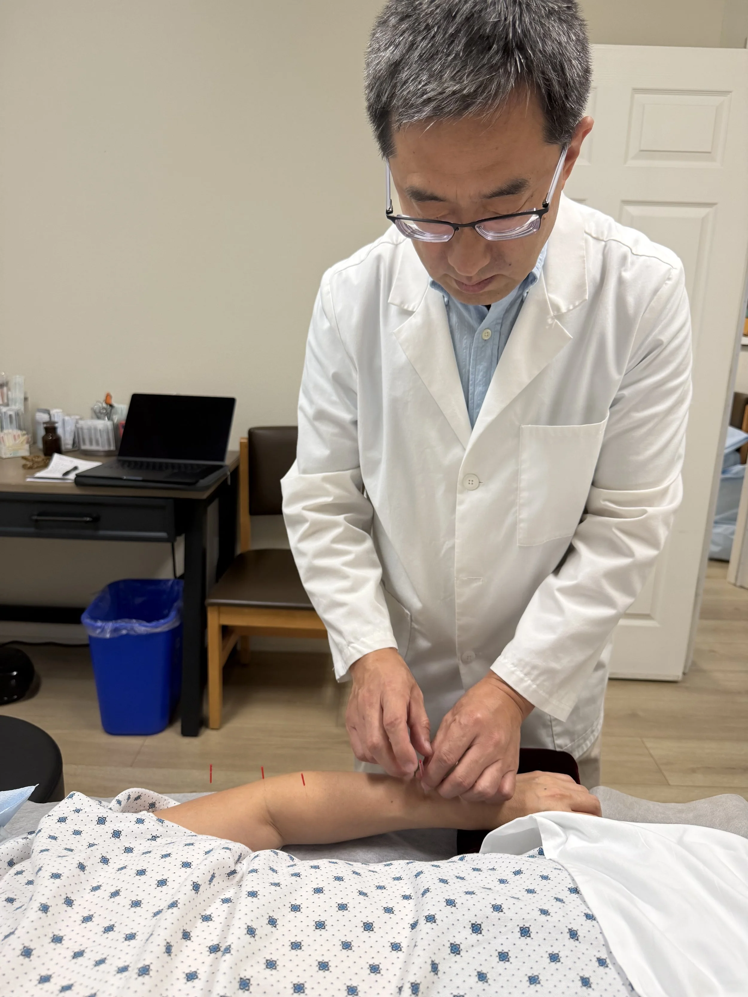 A healthcare professional in a white coat performing acupuncture on a patient's arm in a clinical setting.