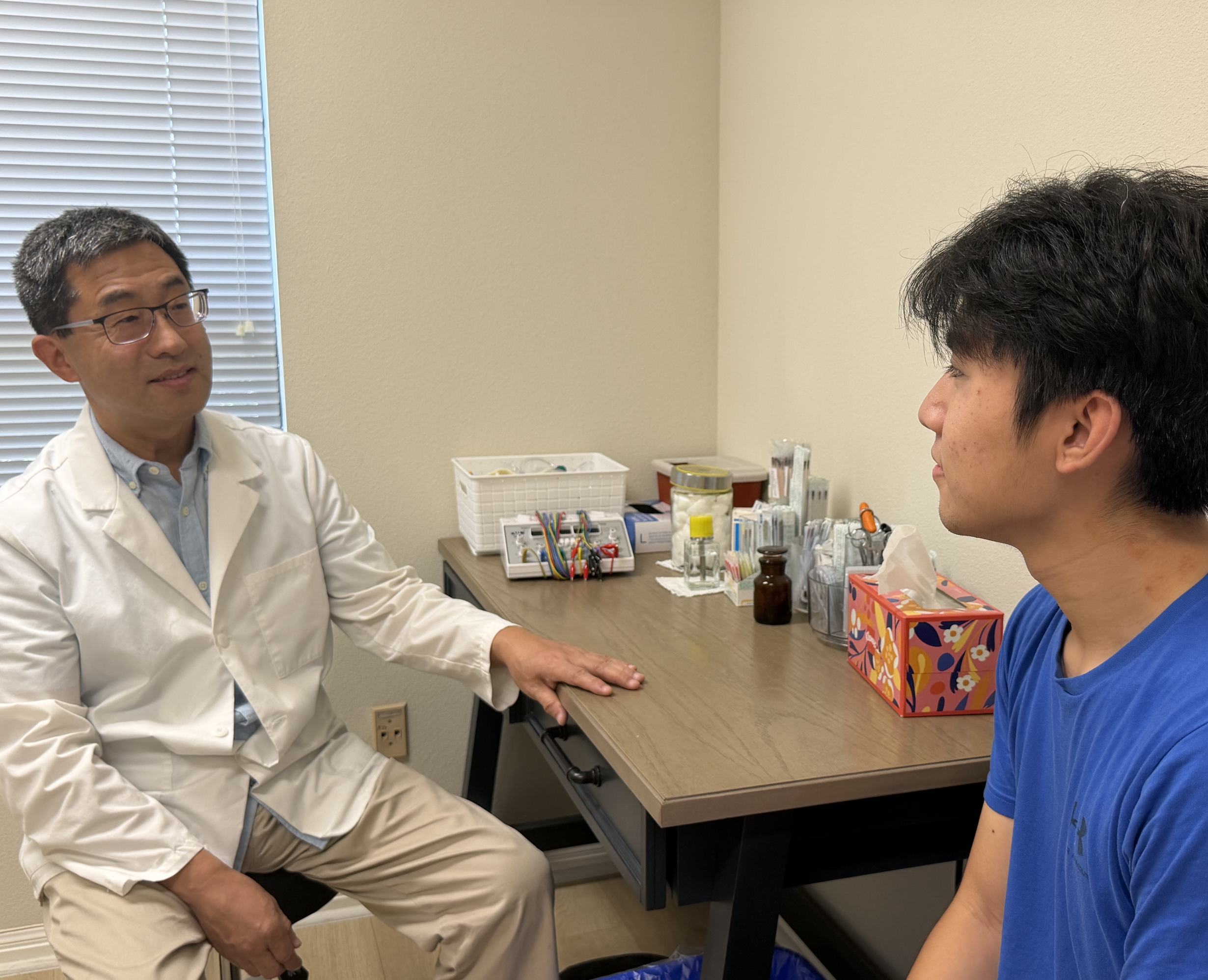 A doctor in a white coat and glasses sitting in a medical office talking to a patient, a young man with dark hair and a blue shirt, who is seated across from him at a desk.