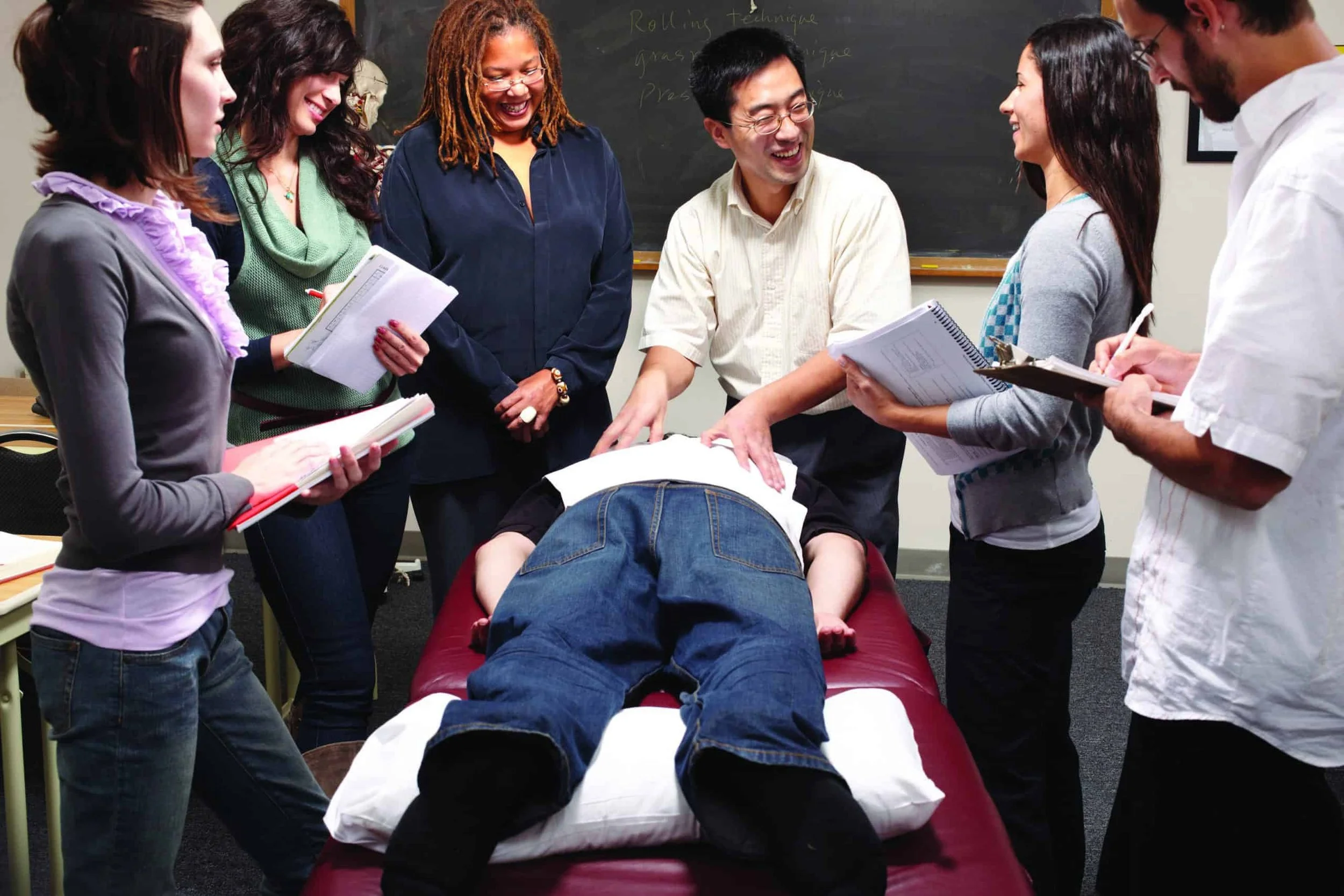 A group of seven people are gathered around a person lying face down on a padded table in a classroom or training room. They appear to be practicing or demonstrating first aid or emergency medical procedures. The people are smiling and seem to be engaged in the activity.