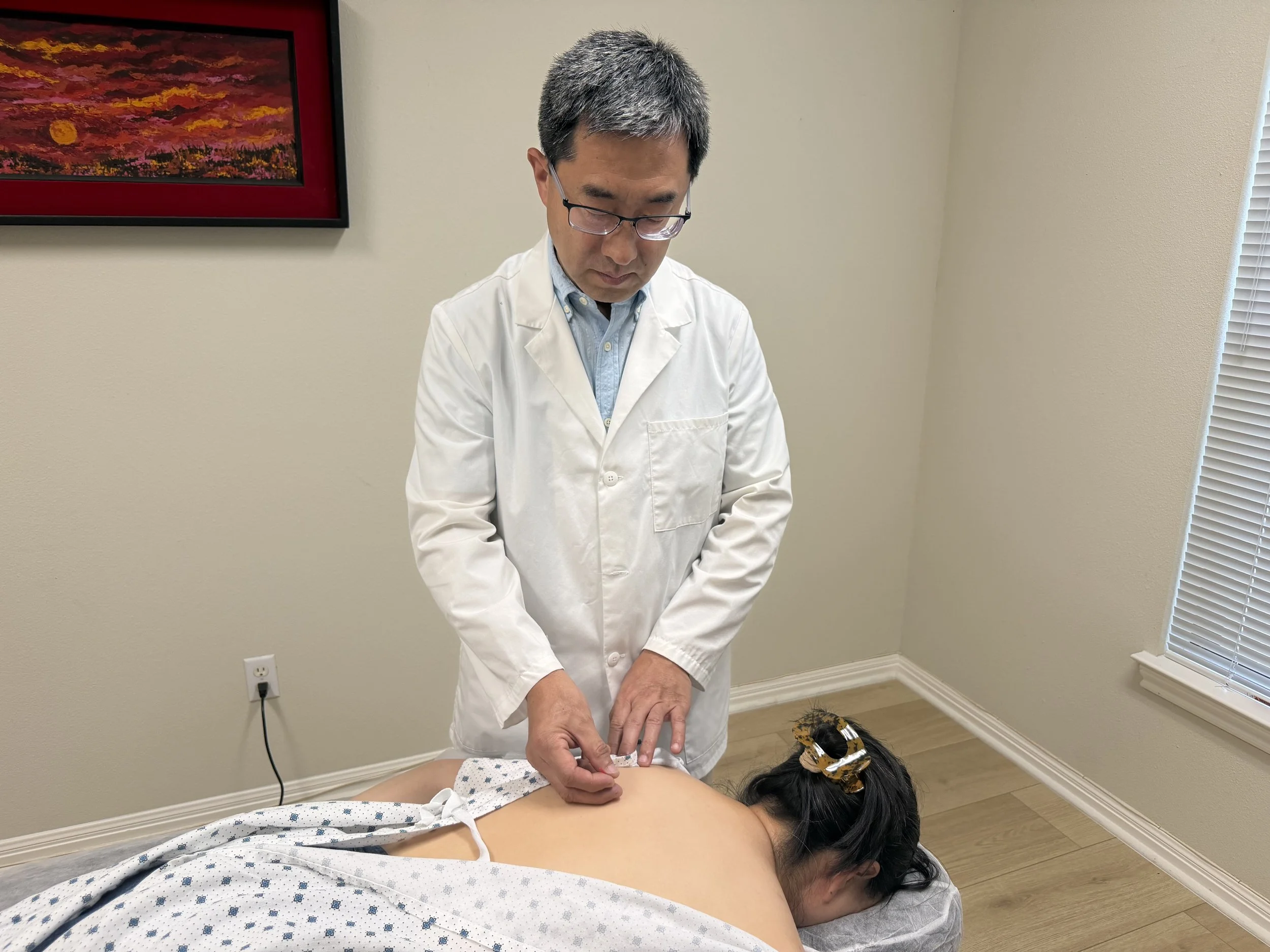 A healthcare professional wearing glasses and a white coat performs acupuncture on a patient lying face down on an examination table in a medical clinic room.