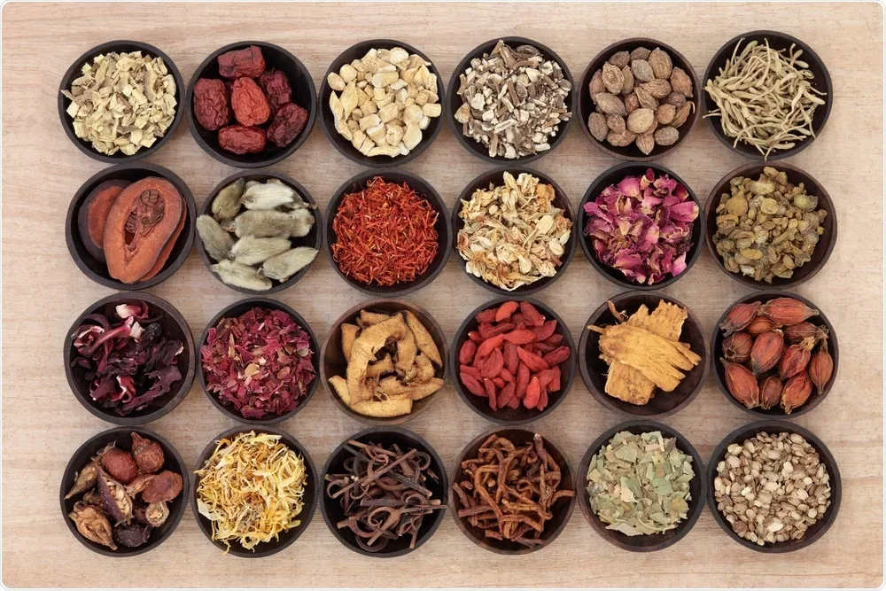 Arrangement of twenty small black bowls containing various dried herbs, spices, and botanicals on a wooden surface.