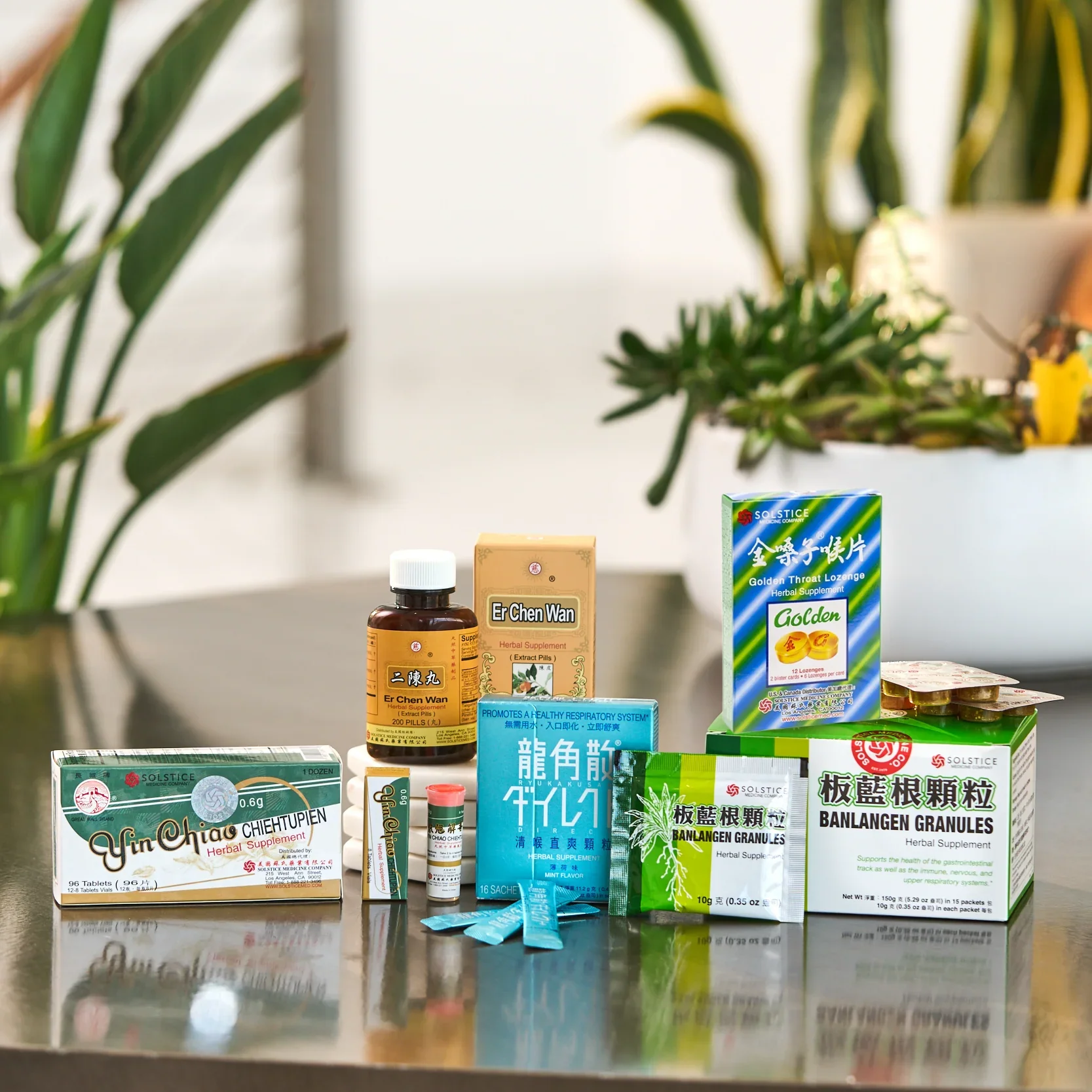 Collection of herbal supplement boxes and bottles on a reflective surface with plants in the background.