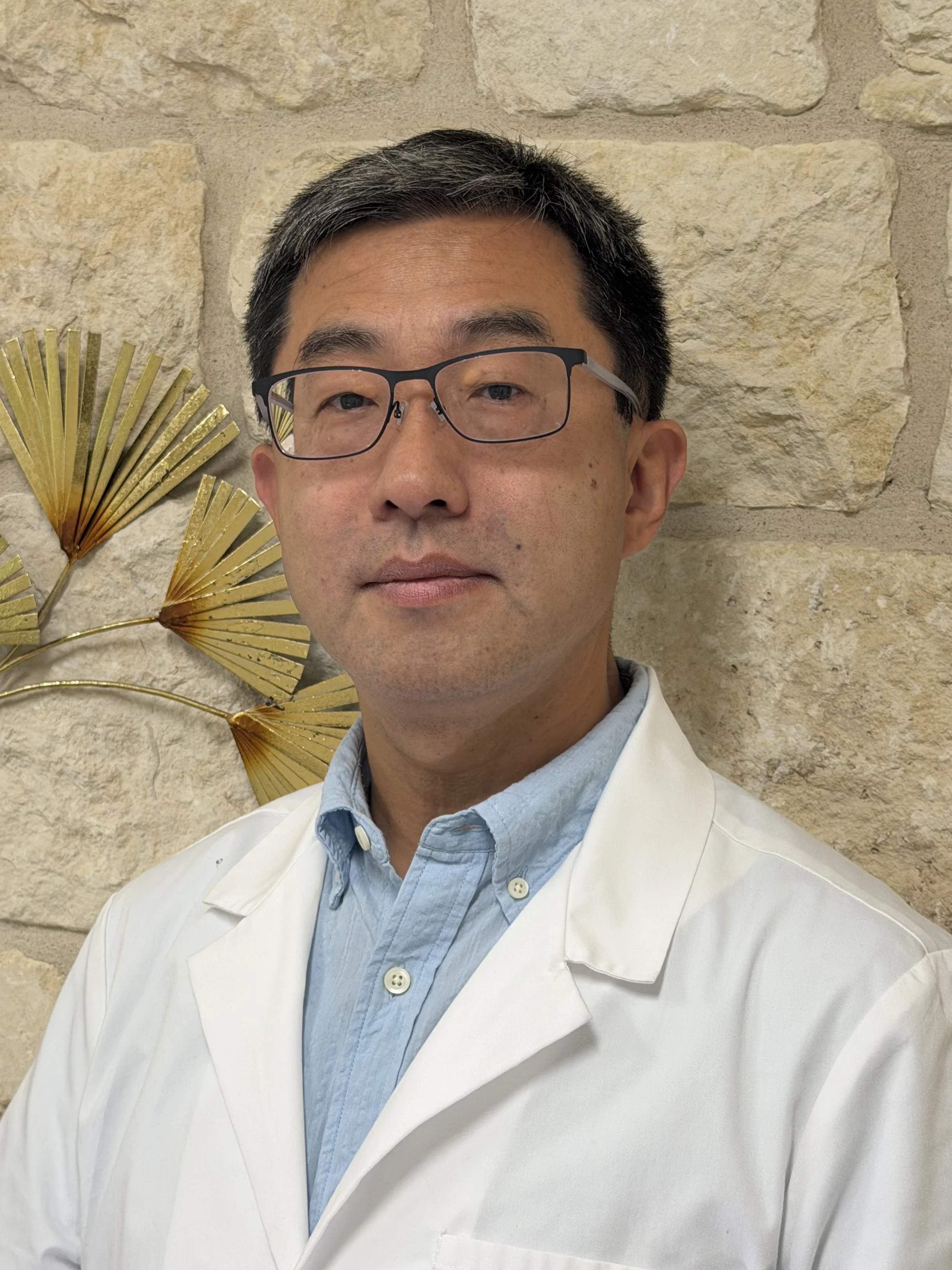 A man wearing glasses, a light blue shirt, and a white coat standing in front of a stone wall with gold-colored decorative palm leaves.